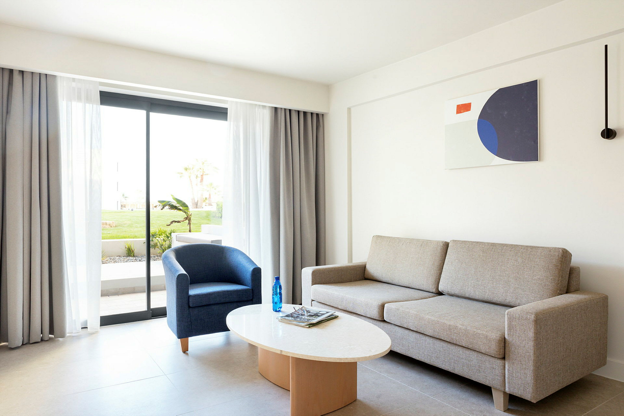 Moderne og lyst opholdsområde med sofa, lænestol og et sofabord. Glasdør til udendørsområde med grønne omgivelser. (AI-genereret tekst)