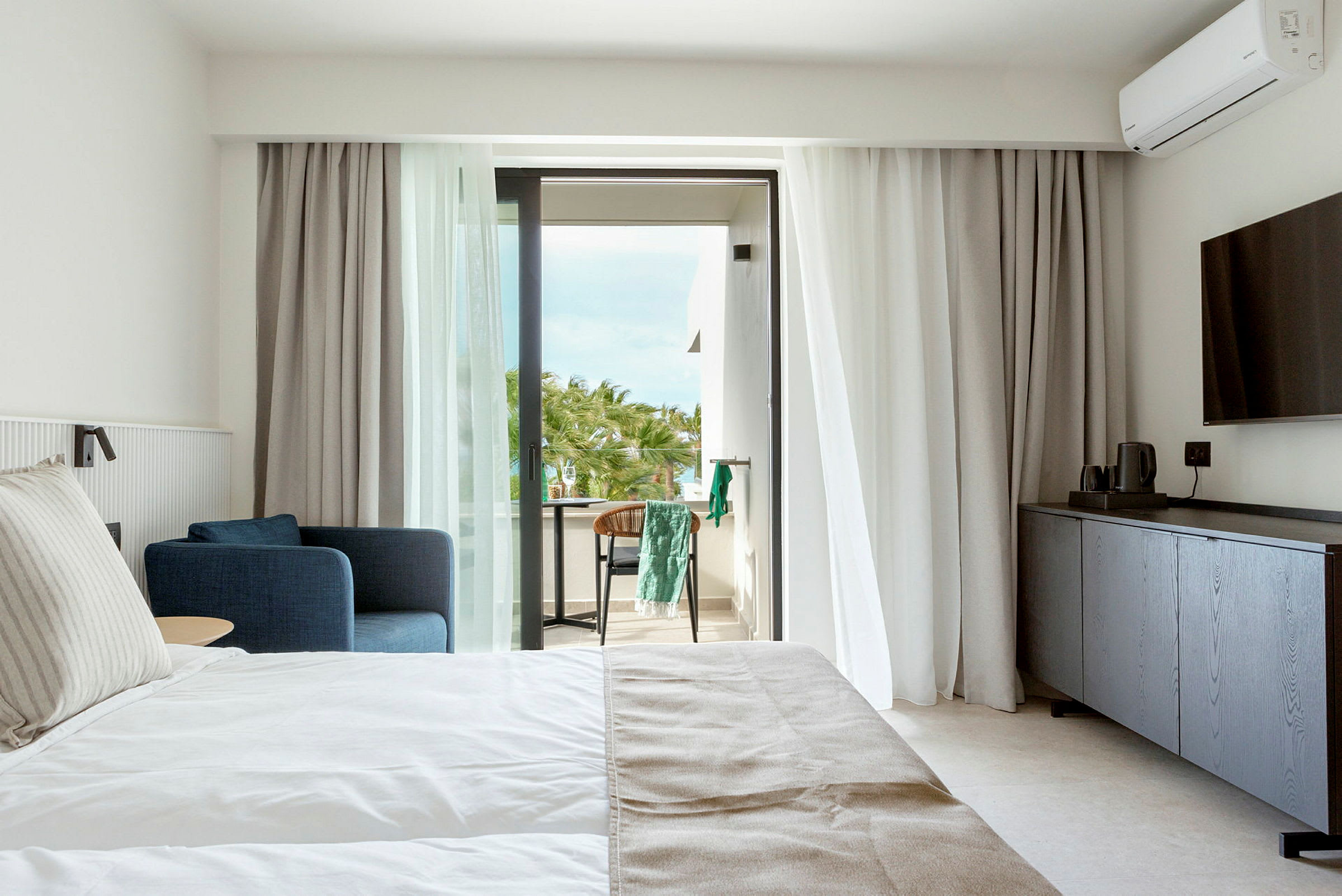 Hotelværelse med seng, blå lænestol og balkon med udsigt til palmer. (AI-genereret tekst)