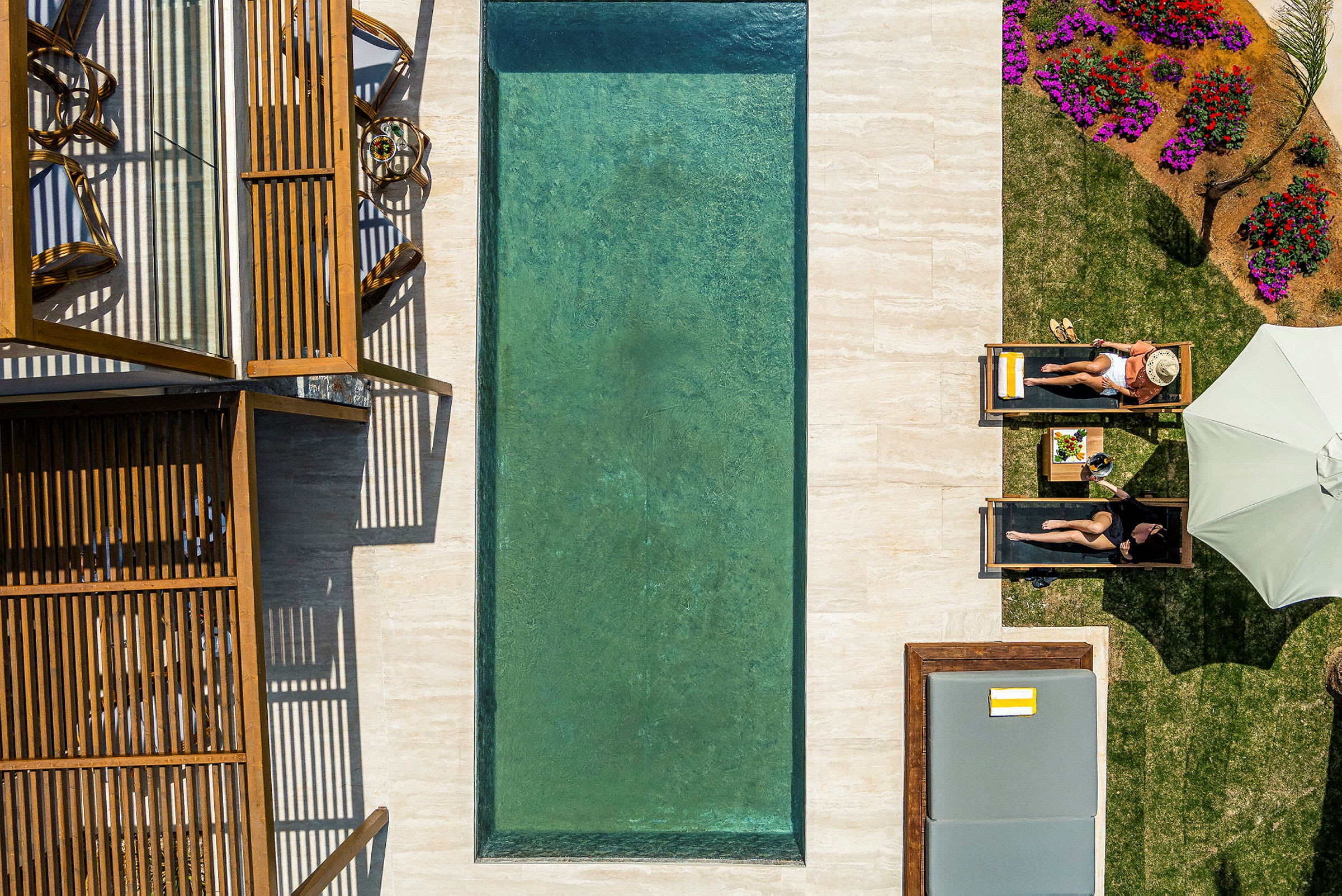 En pool med grønt vand omgivet af solsenge, parasoller og blomsterbede. To personer ligger på solsenge ved siden af poolen. (AI-genereret tekst)