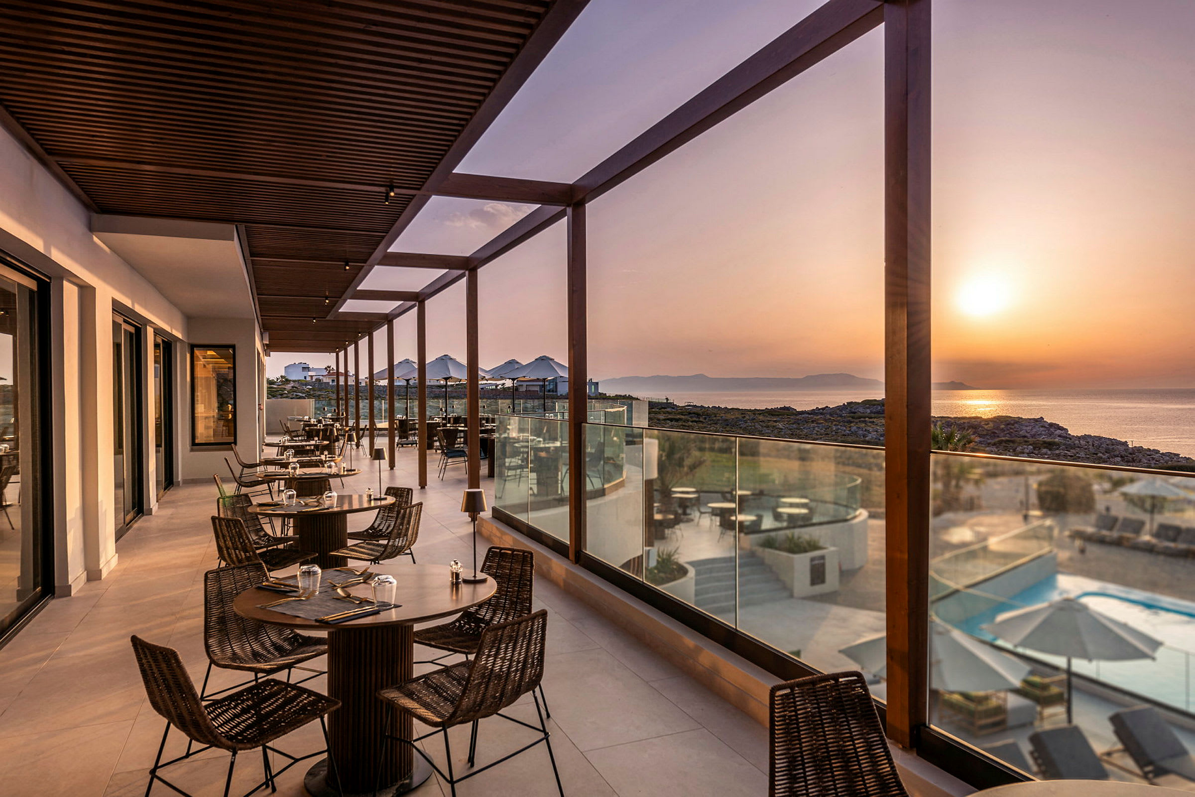 En terrasse med borde og stole, omgivet af glasrækværk, med udsigt over havet og solnedgangen. (AI-genereret tekst)