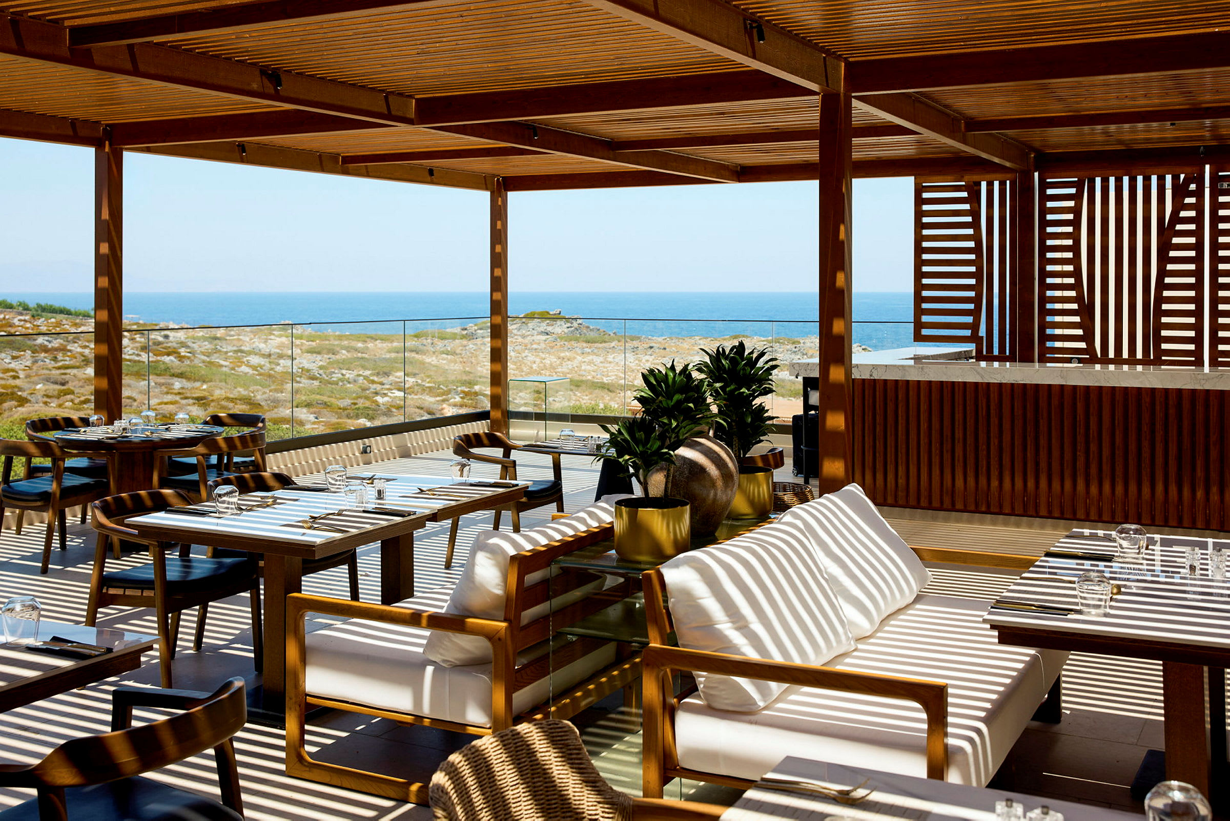 En udendørs restaurant med en træpergola, borde og stole samt havudsigt i baggrunden. (AI-genereret tekst)