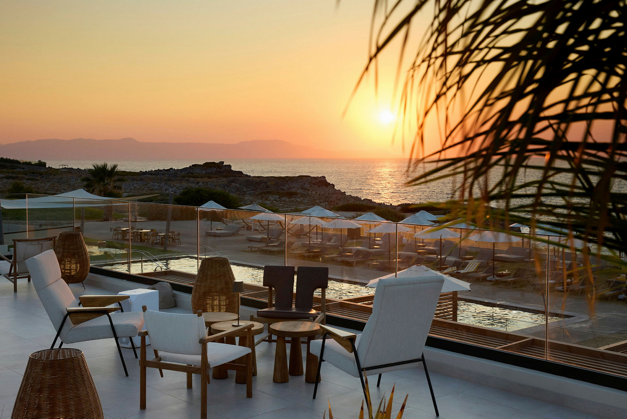 Terrasse med siddepladser i forgrunden, med udsigt over en strand med liggestole og parasoller ved solnedgang. (AI-genereret tekst)
