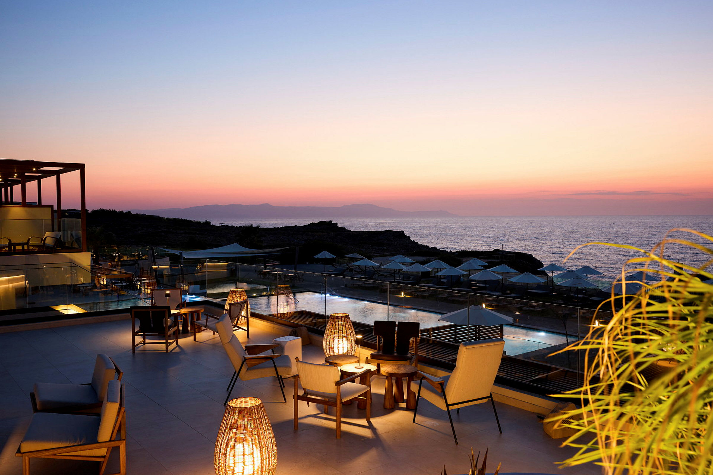 Udsigt over en oplyst terrasse med møbler ved et poolområde, i baggrunden et hav og en smuk solnedgang. (AI-genereret tekst)