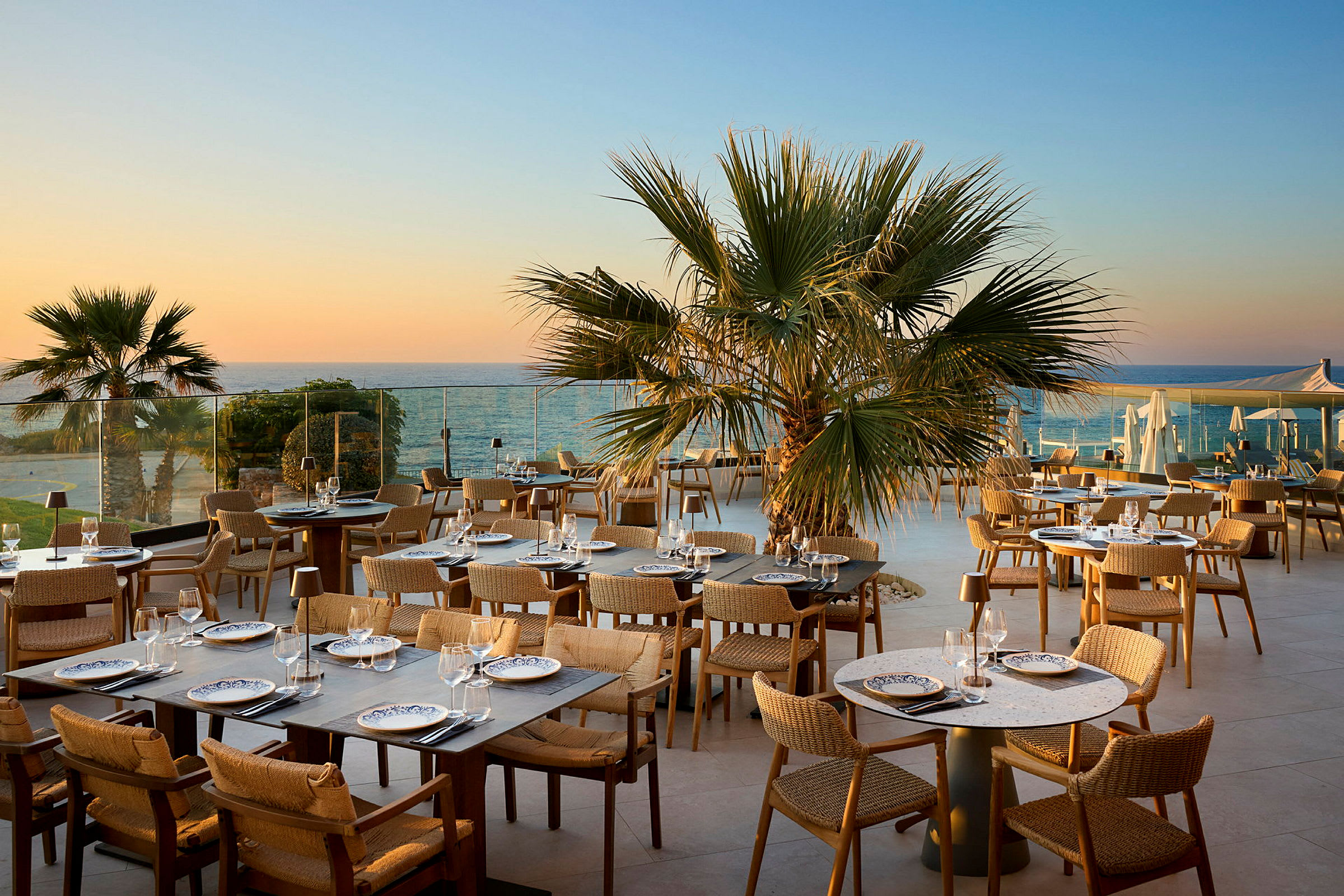 En udendørs restaurant med borde og stole, omgivet af palmer og med udsigt til havet i solnedgangen. (AI-genereret tekst)