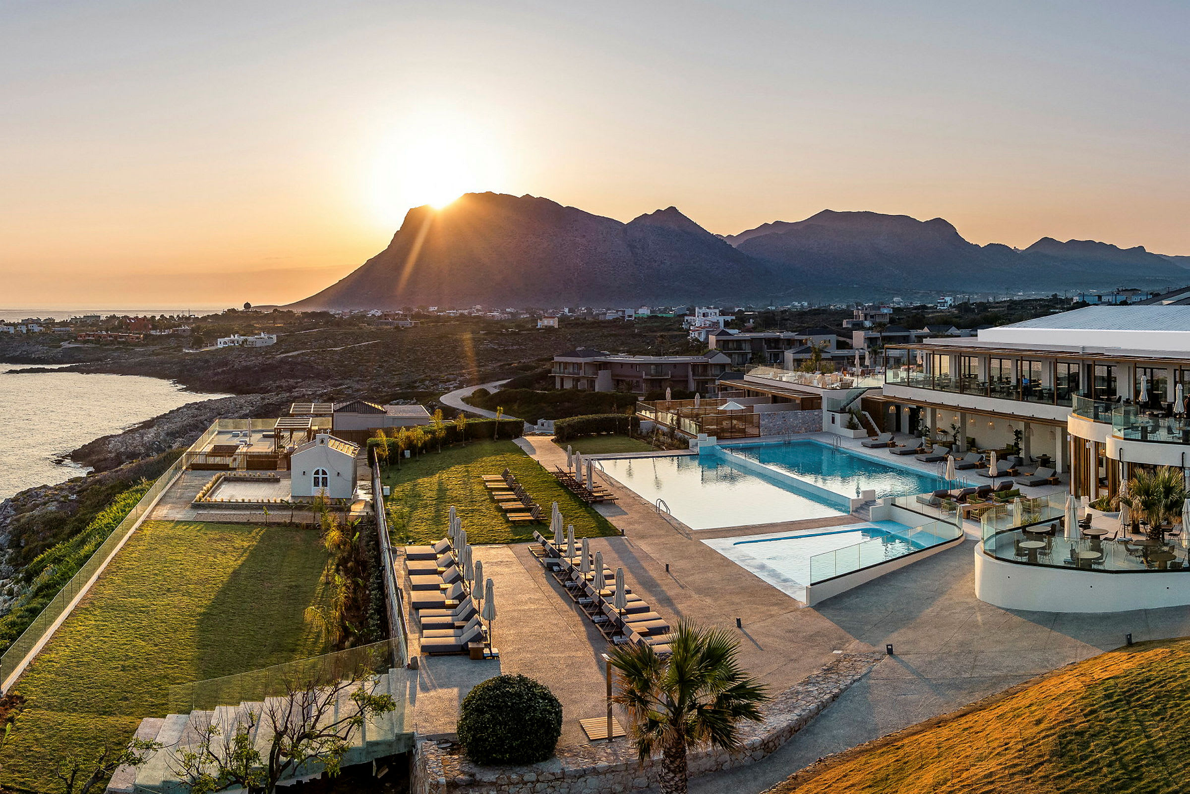 Solnedgang over hotelområde med pool, grønne områder og omkringliggende bjerglandskab. (AI-genereret tekst)
