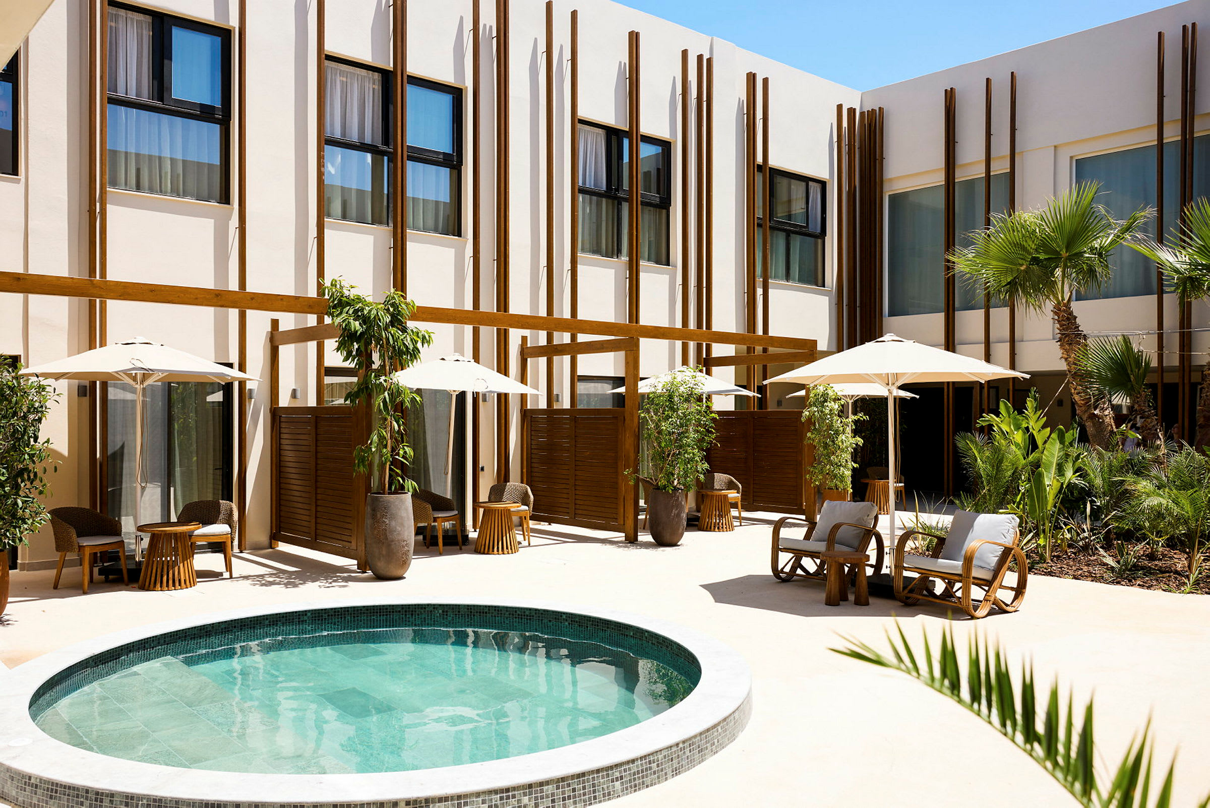 Et moderne hotelområde med en lille rund pool omgivet af beplantning, liggestole og parasoller. (AI-genereret tekst)