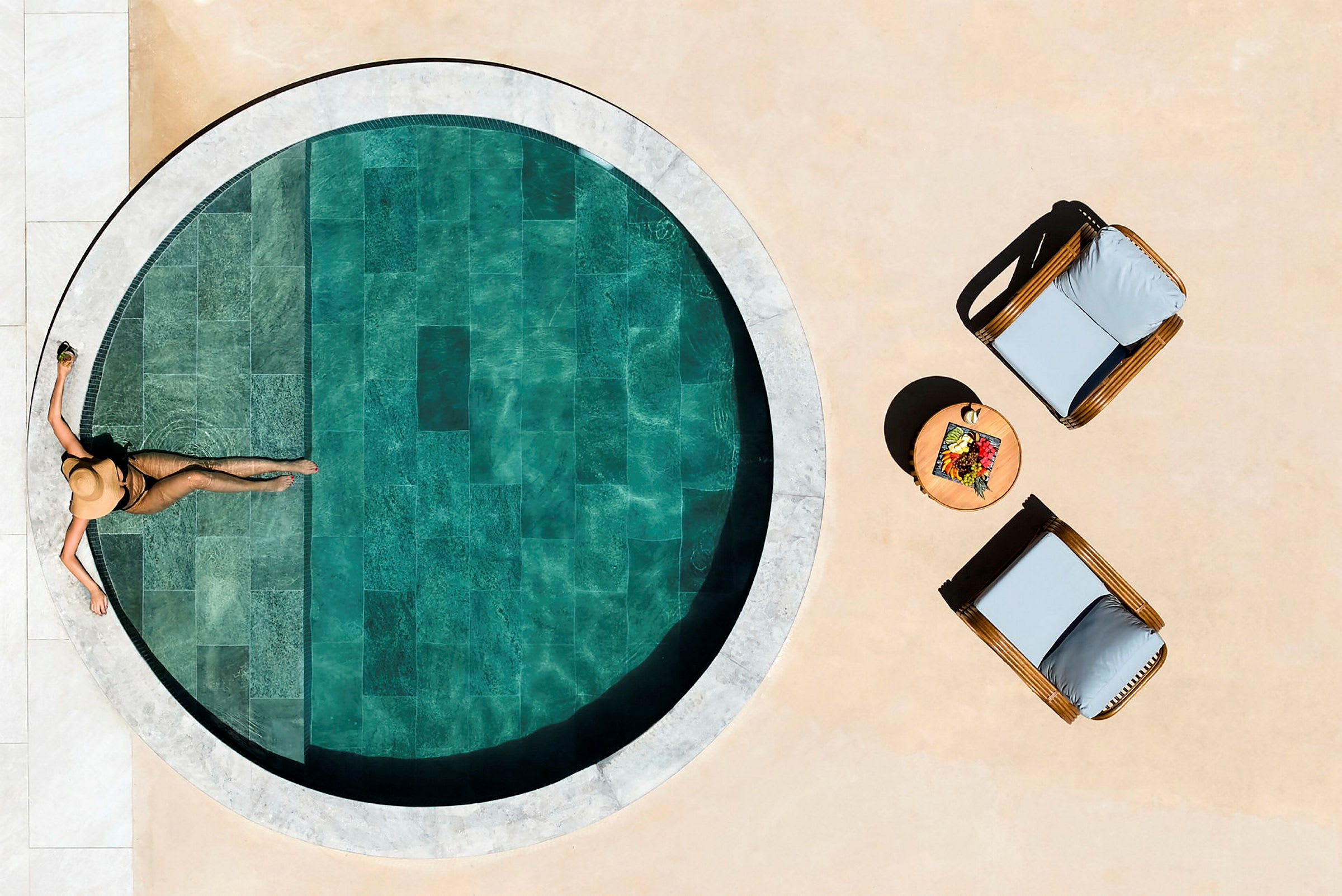 En person slapper af i solen ved en lille, rund pool, med to solsenge og et bord med frugt ved siden. (AI-genereret tekst)