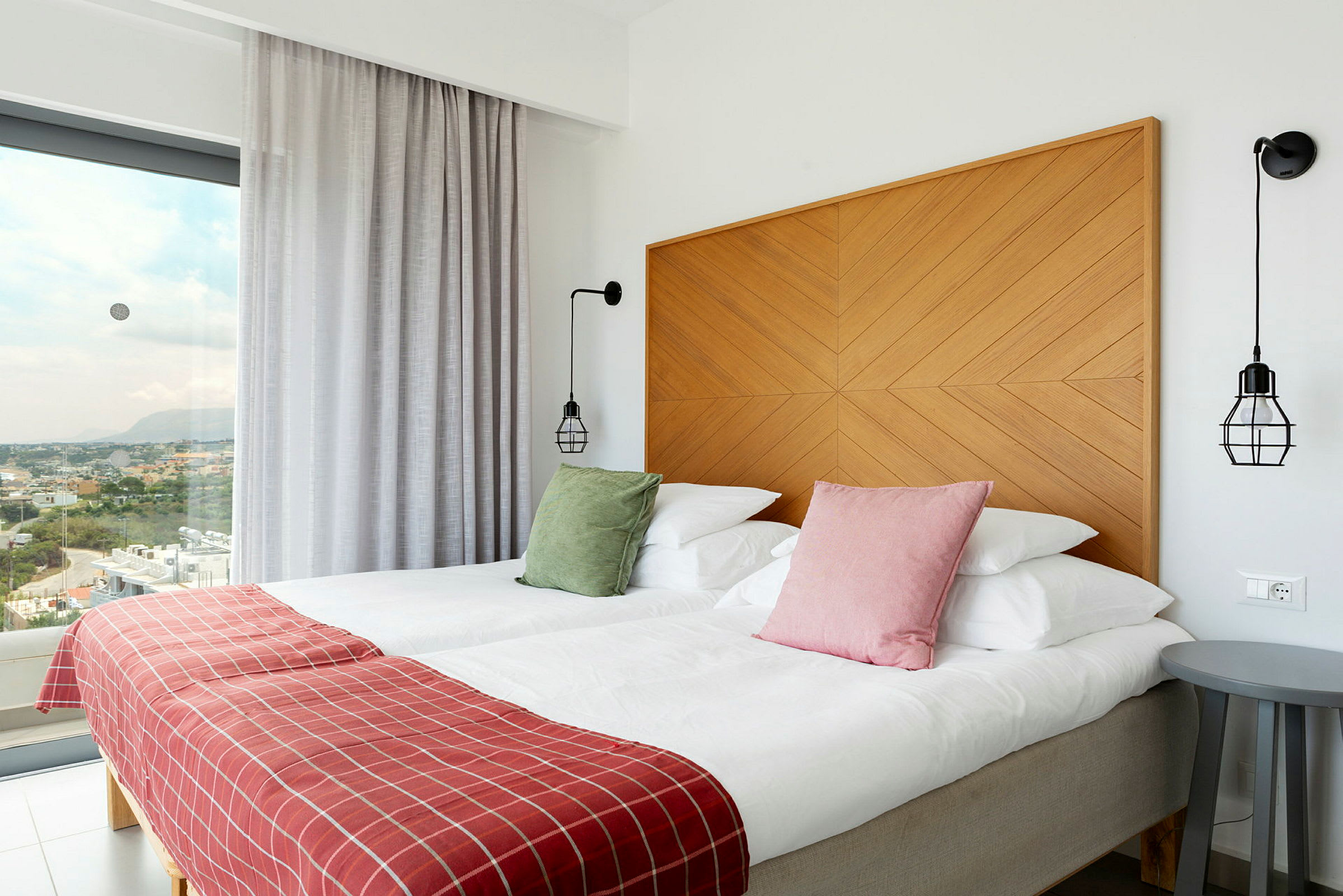 Hotelværelse med to senge, farverige puder, rødt sengetæppe og moderne indretning, med udsigt gennem store vinduer. (AI-genereret tekst)