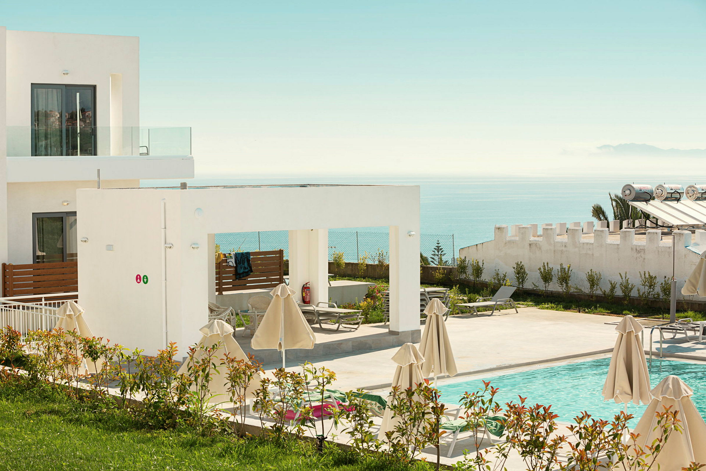 Billede af et hotel med moderne arkitektur, en pool og solsenge omgivet af parasoller samt udsigt over havet. (AI-genereret tekst)