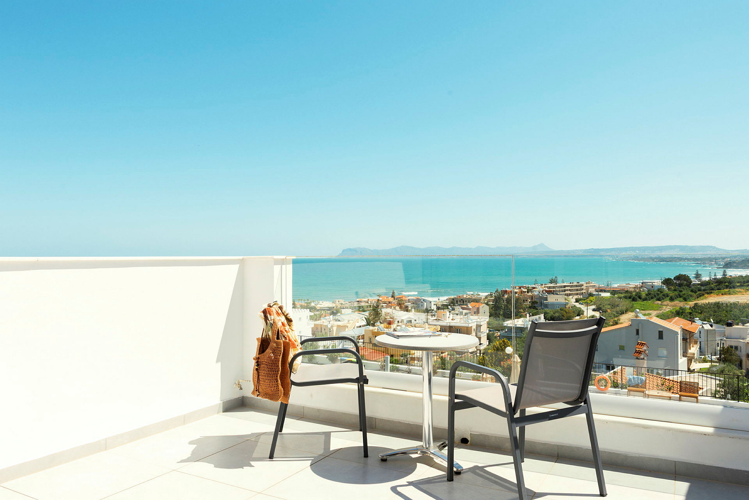 Terrasse med to stole og et bord foran glasrækværk med udsigt over hav og byer. (AI-genereret tekst)