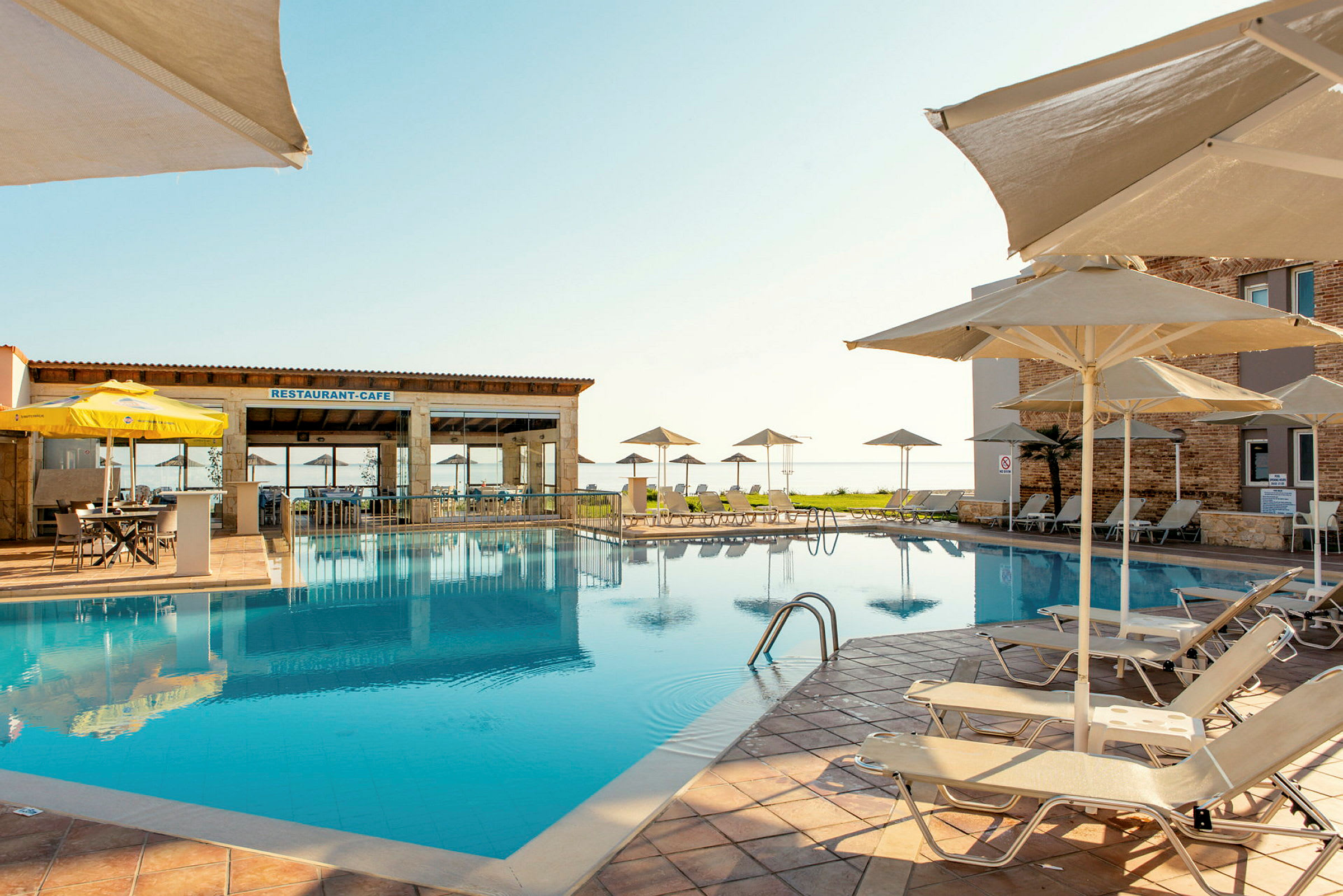 En udendørs pool med liggestole og parasoller ved et hotel. En restaurantbygning ses i baggrunden. (AI-genereret tekst)
