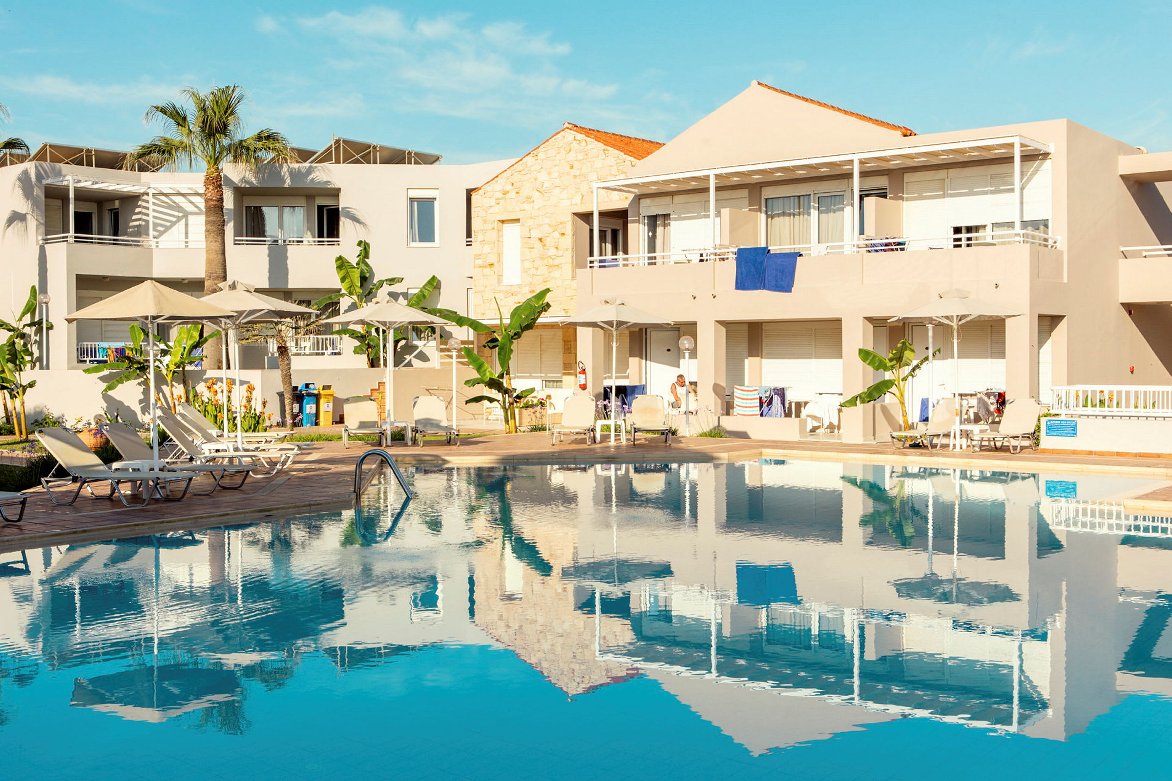 Lejlighedshotel med pool og liggestole under parasoller. (AI-genereret tekst)
