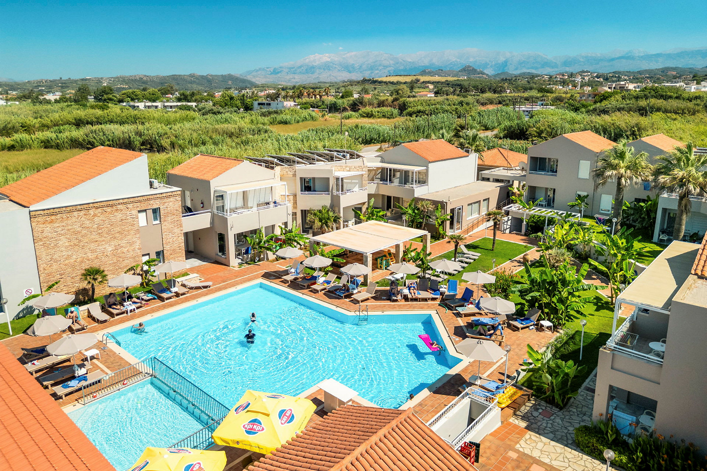 Hotelområde med en pool omgivet af solsenge og parasoller, med grønne omgivelser og bygninger i baggrunden. (AI-genereret tekst)