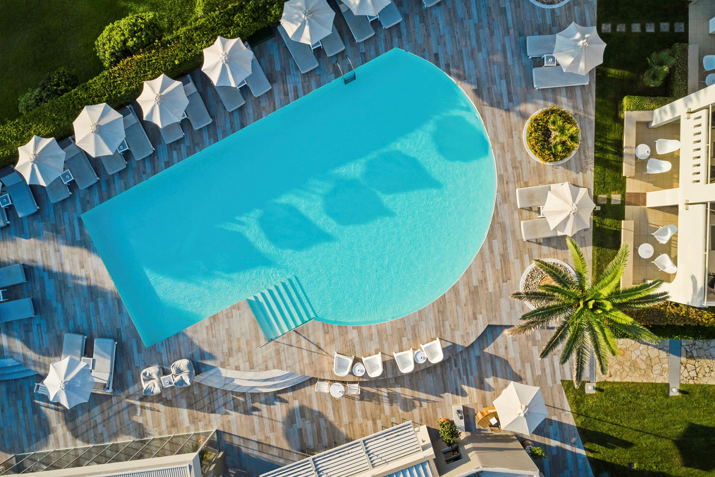 Luftfoto af en pool, liggestole og parasoller samt græsareal på et feriested. (AI-genereret tekst)