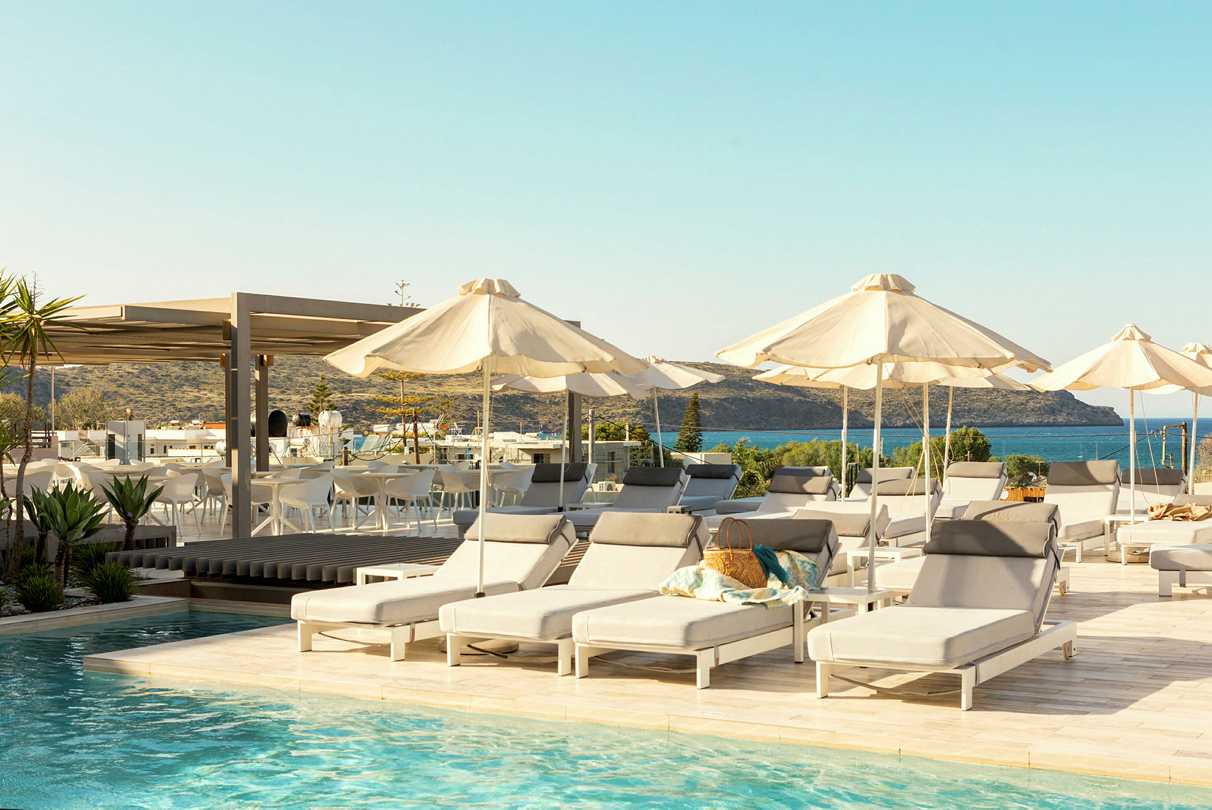 Billedet viser en pool med liggestole og parasoller omkring. I baggrunden ses havet og en terrasse med siddepladser. (AI-genereret tekst)