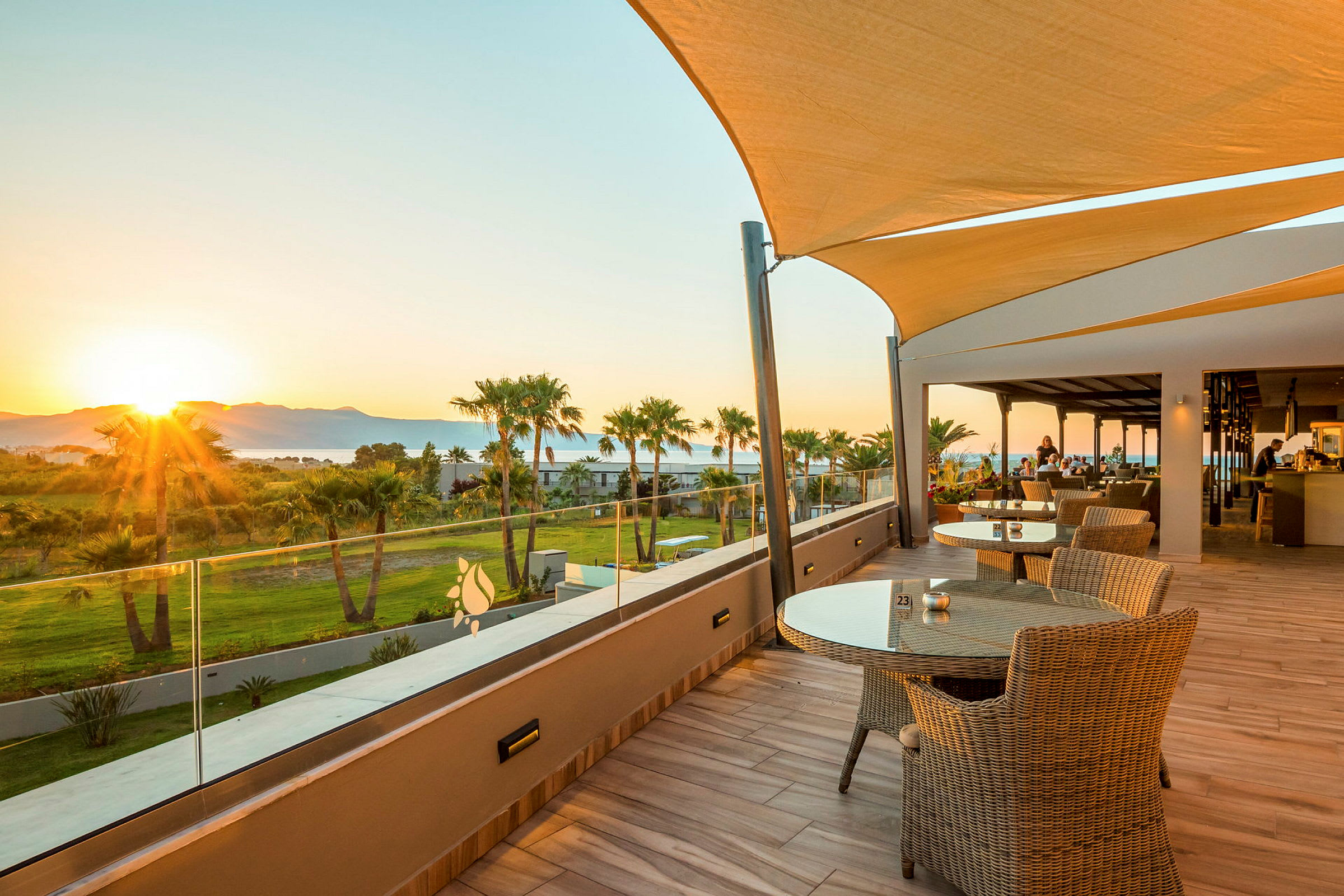 Terrasse med borde og stole samt udsigt over grønne områder og solnedgang. (AI-genereret tekst)