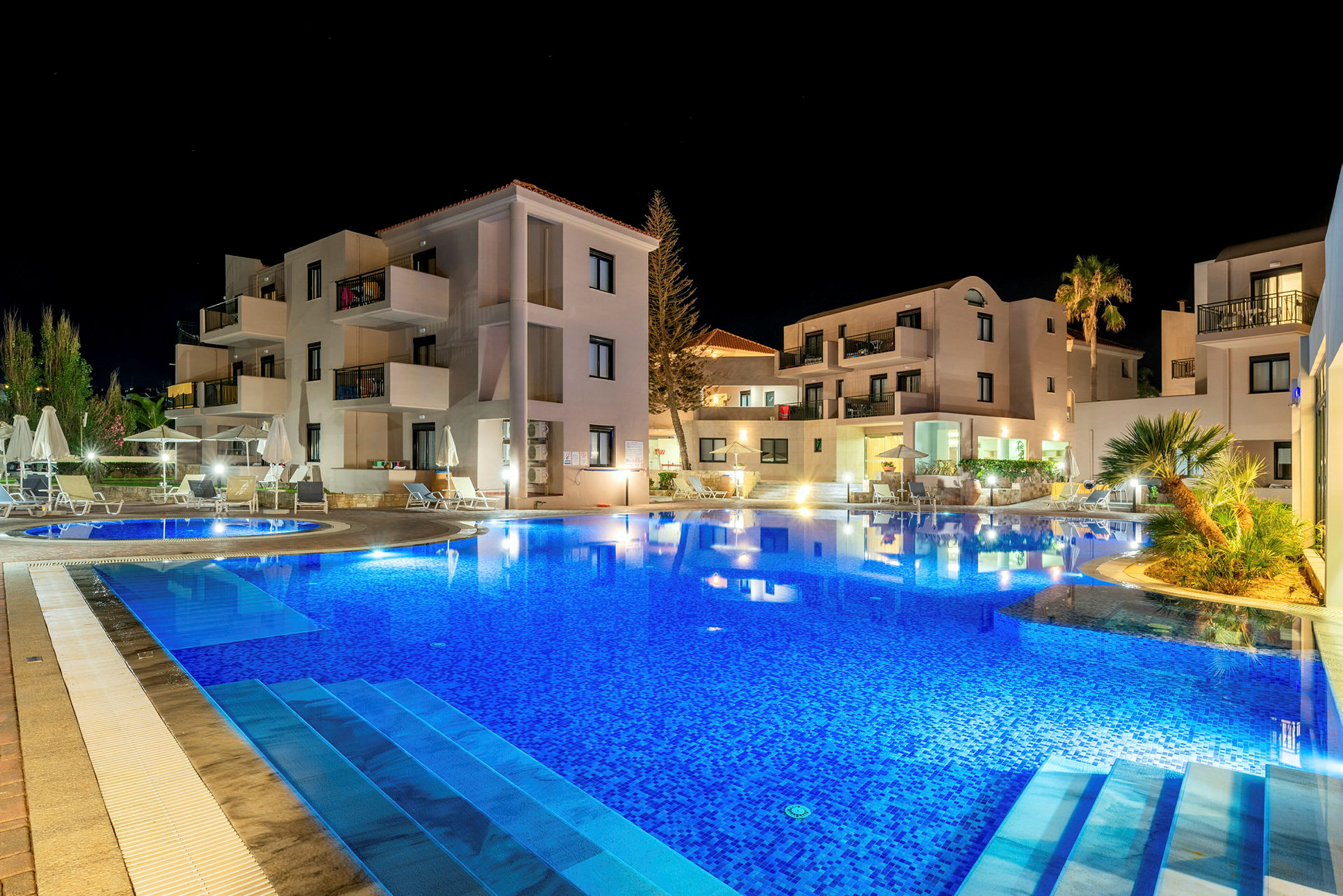 Hotelbygning oplyst om aftenen med en swimmingpool i forgrunden. (AI-genereret tekst)