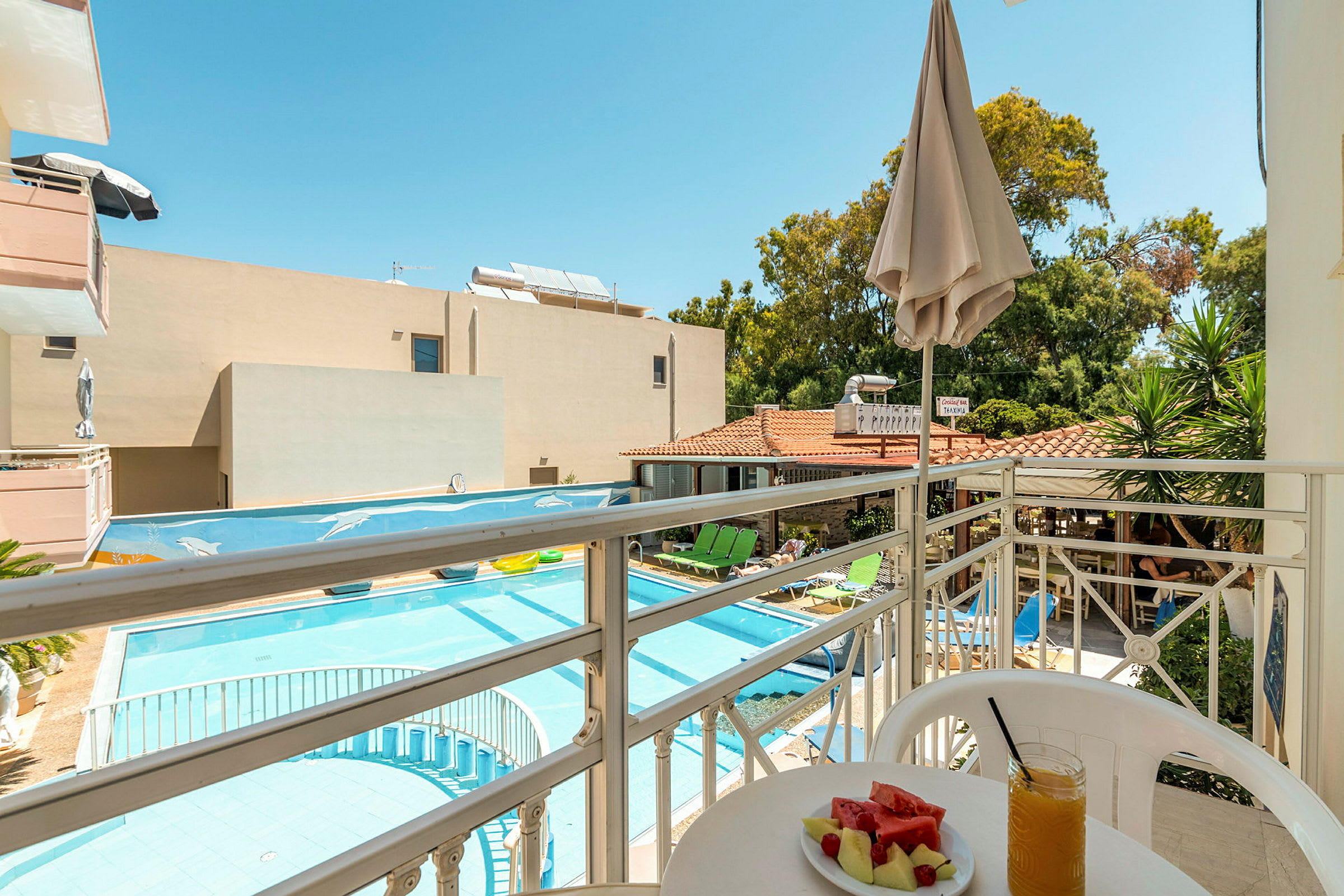 Udsigt fra en balkon mod en pool og hotelområde med liggestole, parasoller og grønne områder. På balkonen står et bord med en tallerken melon og et glas juice. (AI-genereret tekst)