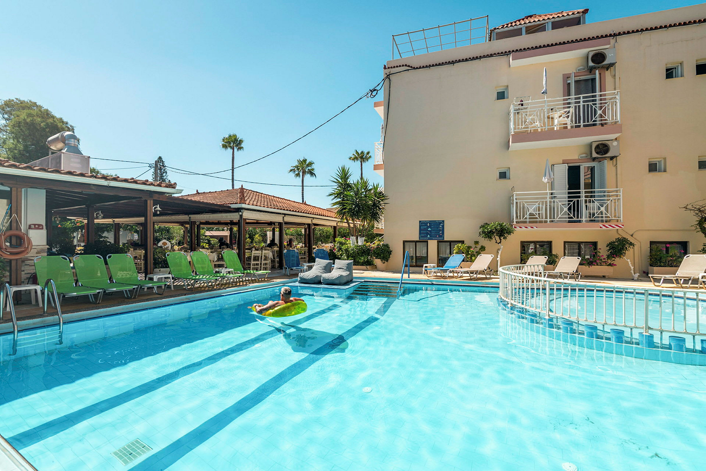 Hotel med liggestole omkring en udendørs pool og en person, der ligger på en oppustelig madras i vandet. (AI-genereret tekst)