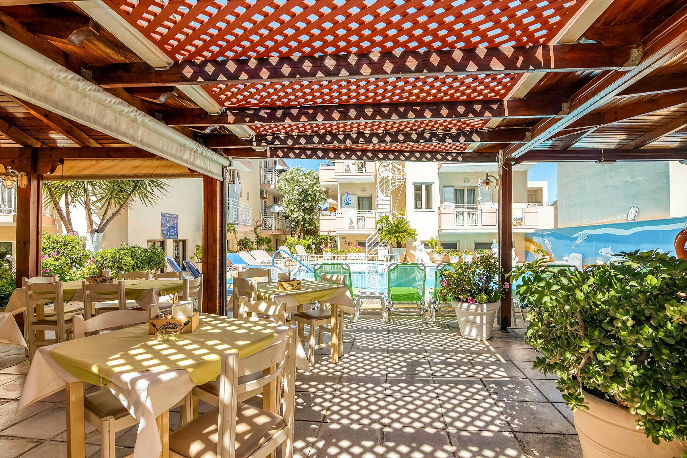 Billede af et udendørs spiseområde med borde og stole under en træpergola, med udsigt til en pool og bygninger i baggrunden. (AI-genereret tekst)