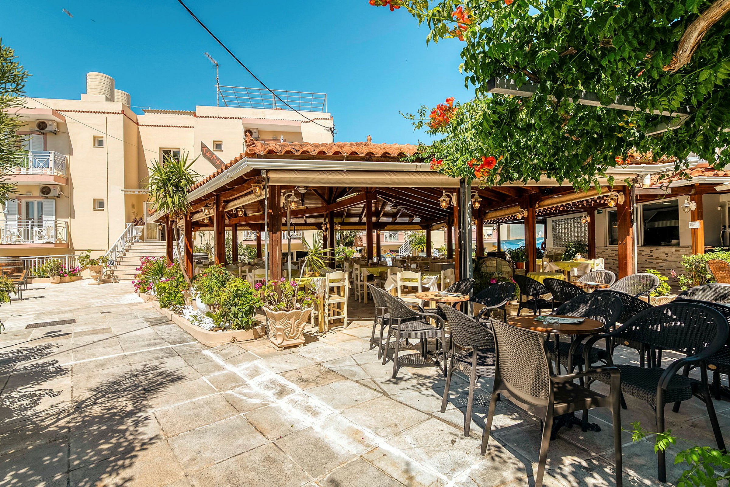 En udendørsrestaurant med borde og stole under en pergola, omgivet af grønne planter. (AI-genereret tekst)