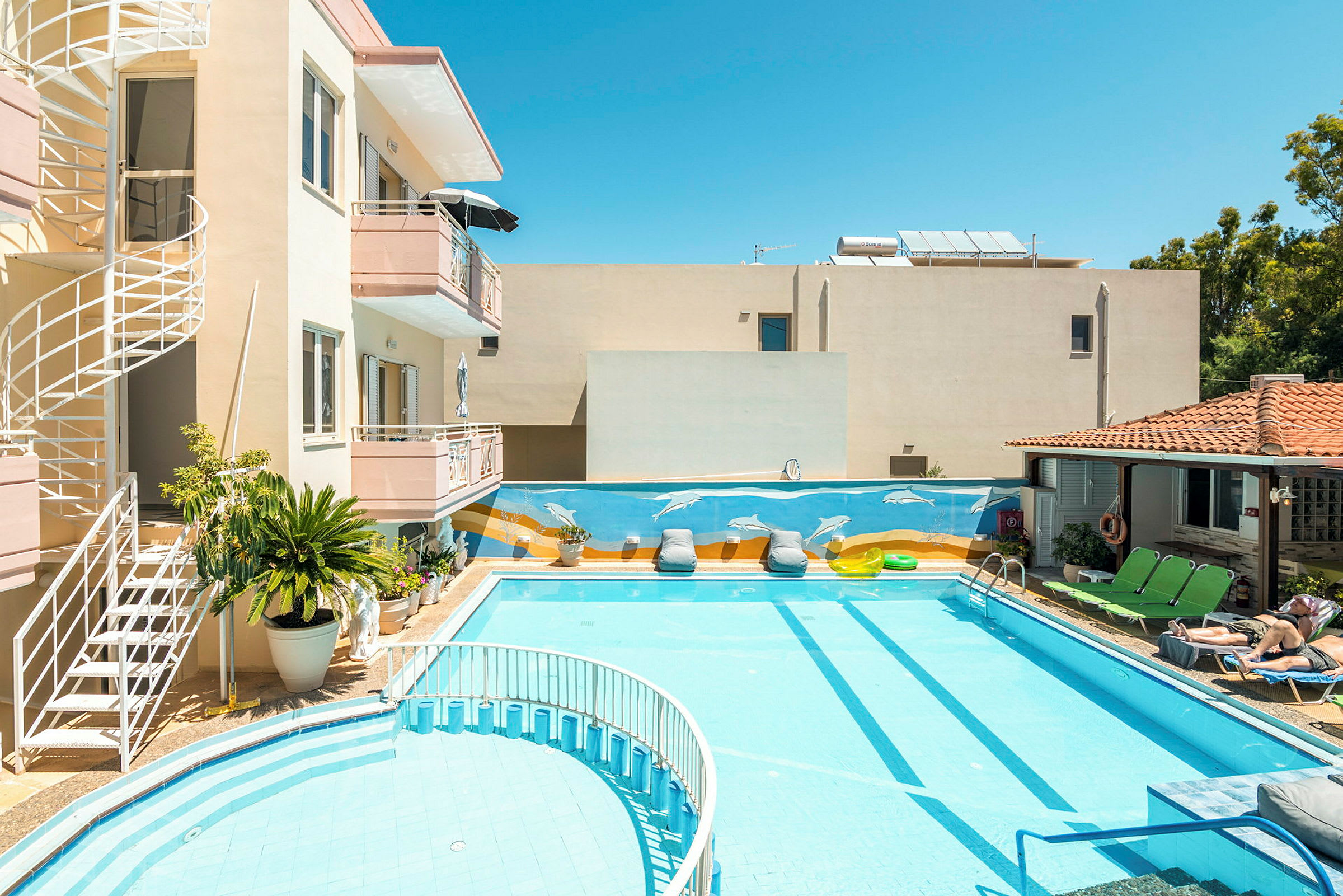 En bygning med altaner og en vindeltrappe ved en swimmingpool. Poolområdet har liggestole og dekorationer, herunder et vægmaleri med delfiner. (AI-genereret tekst)