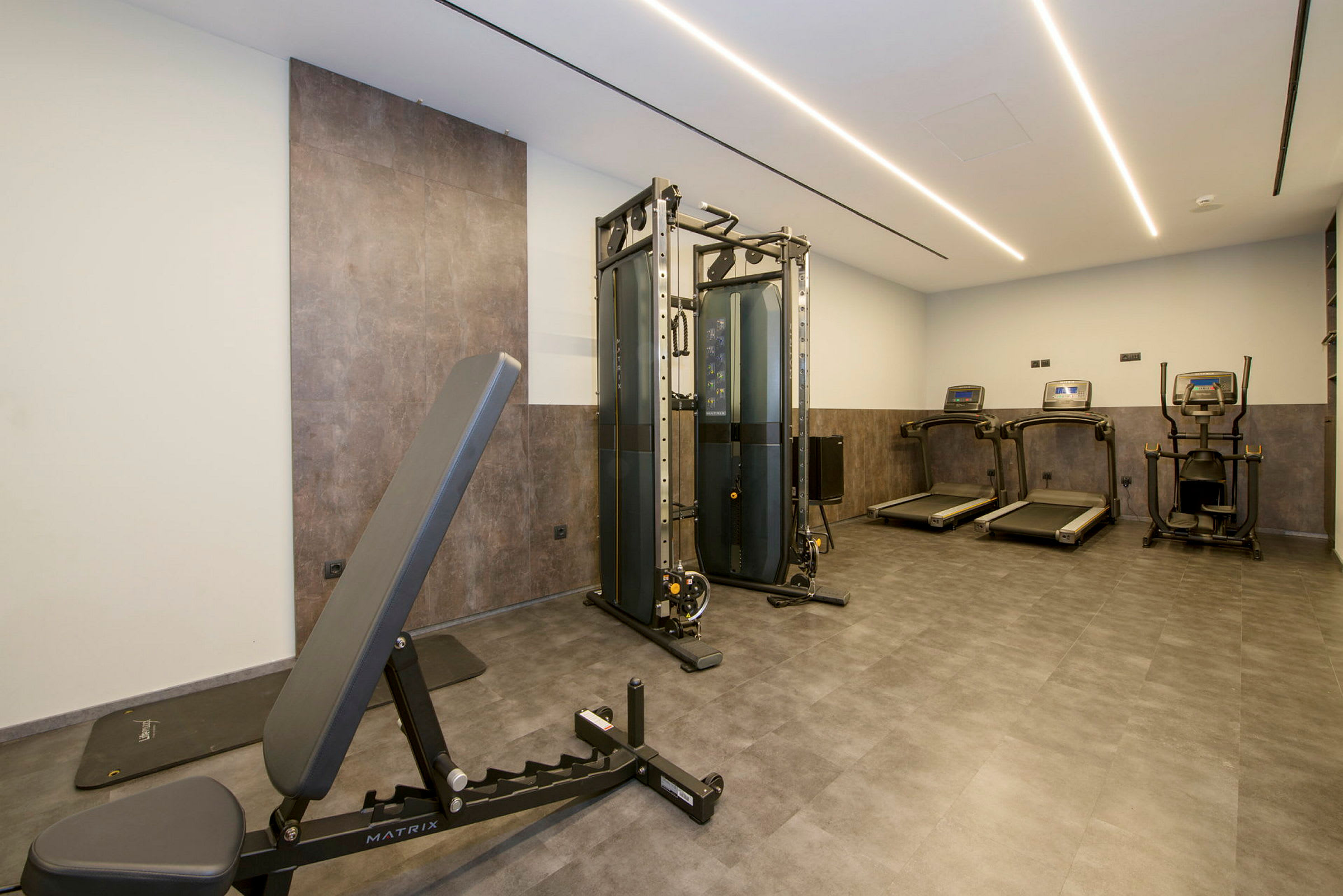 Fitnessudstyr inklusive løbebånd, træningsmaskine og bænk på et gulv med grå fliser. (AI-genereret tekst)