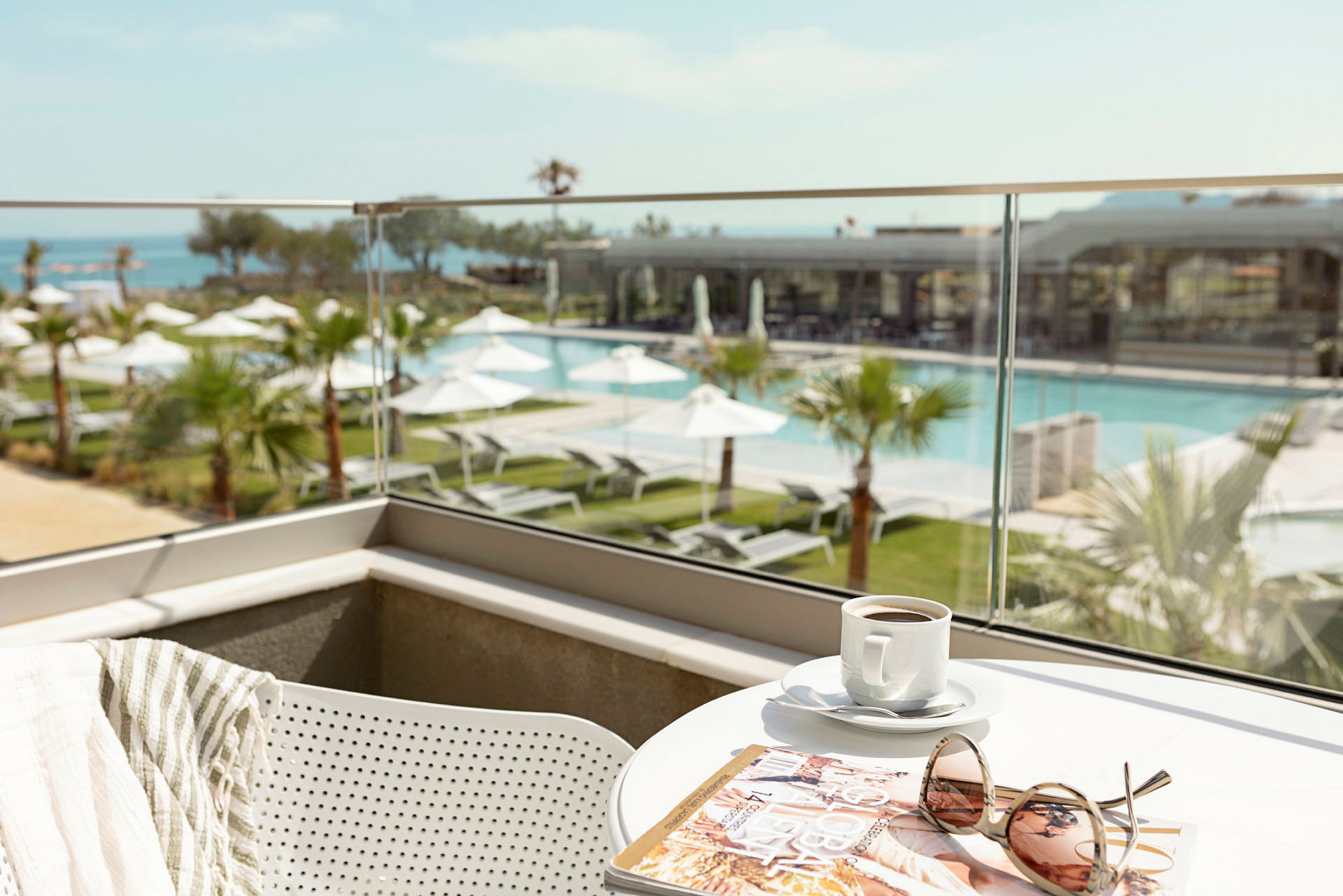 Udsigt over en pool og have fra en balkon med et bord med en kaffekop, et magasin og solbriller. (AI-genereret tekst)