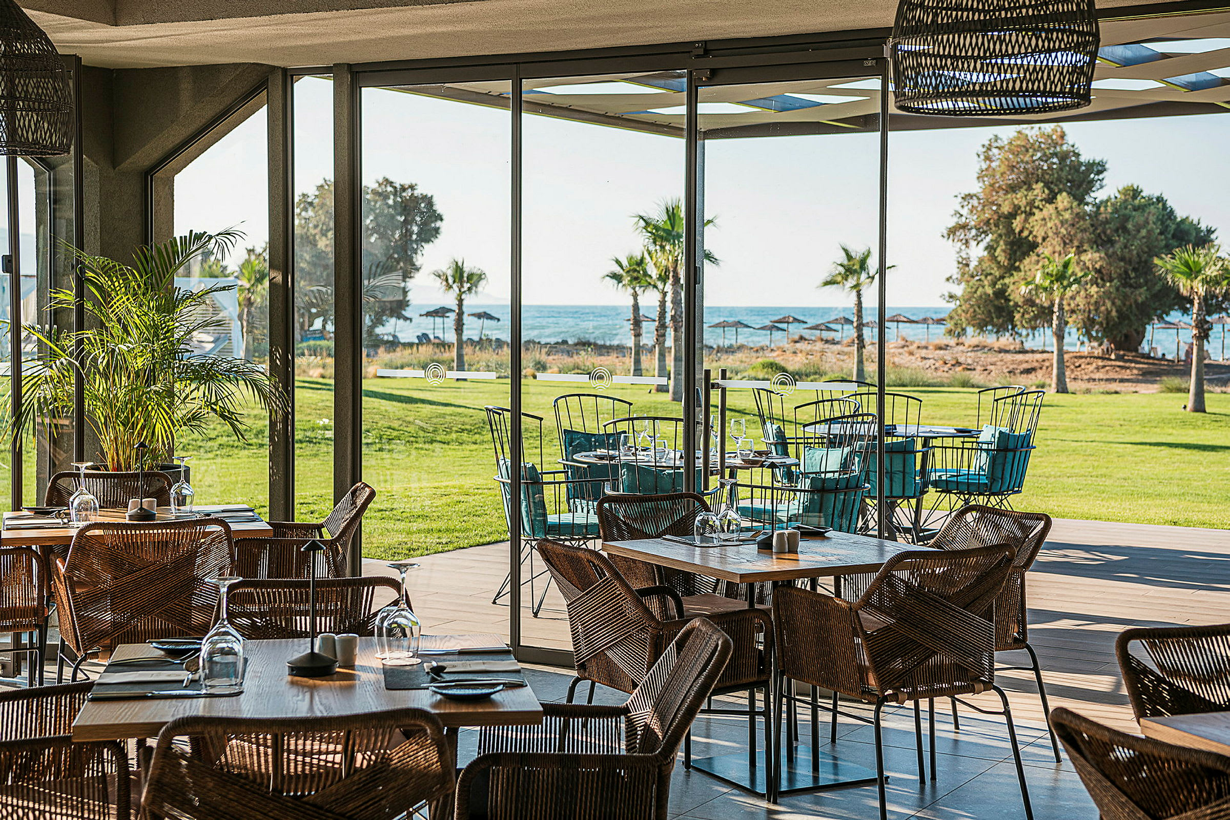 En restaurant med glasvægge og udsigt over en grøn plæne, palmer og havet. (AI-genereret tekst)