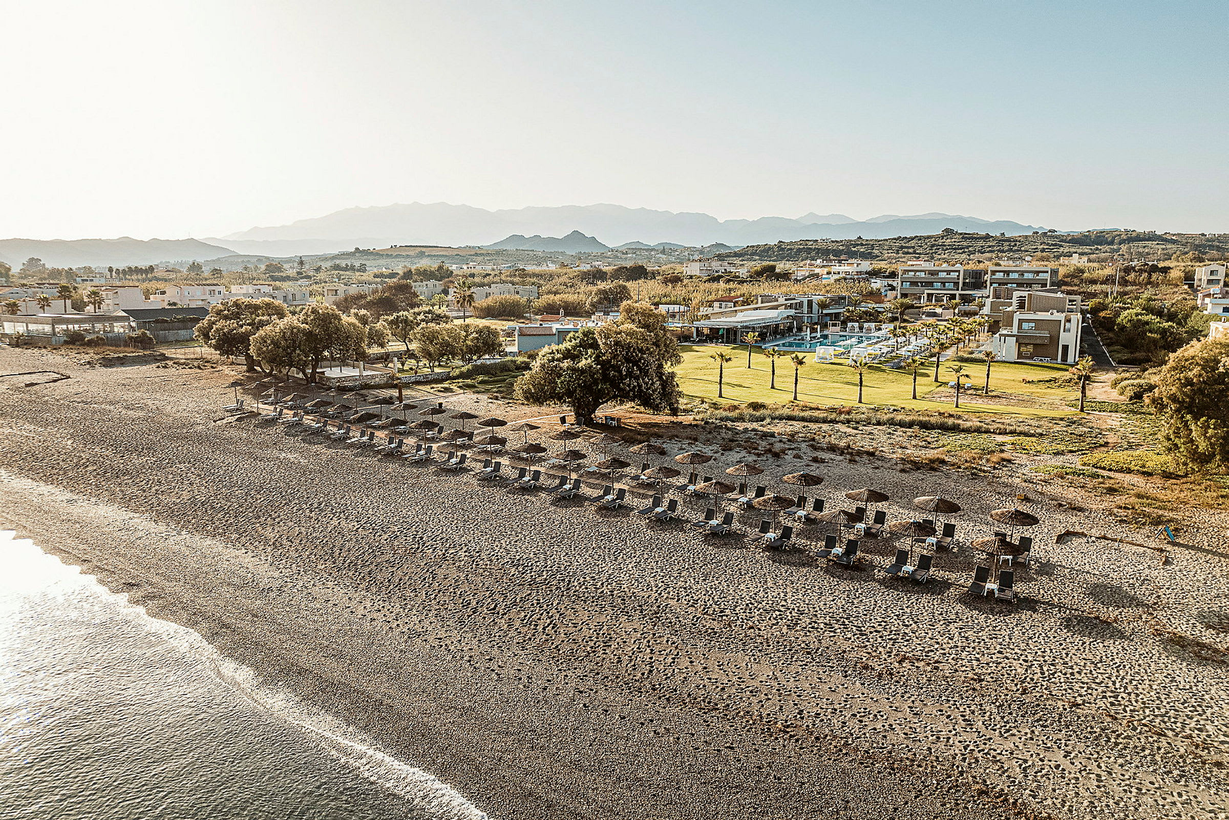 En strand med liggestole og parasoller, omgivet af grønne områder og hotelbygninger i baggrunden. (AI-genereret tekst)