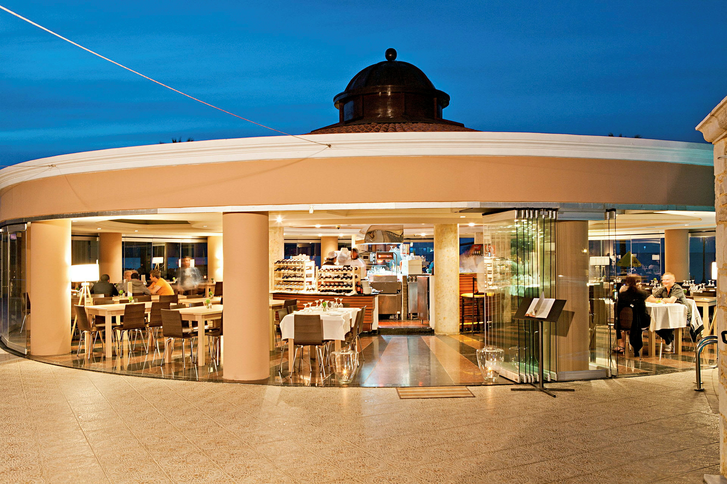 En restaurant med glasvægge og borde placeret indendørs, set fra en udendørsterrasse. (AI-genereret tekst)