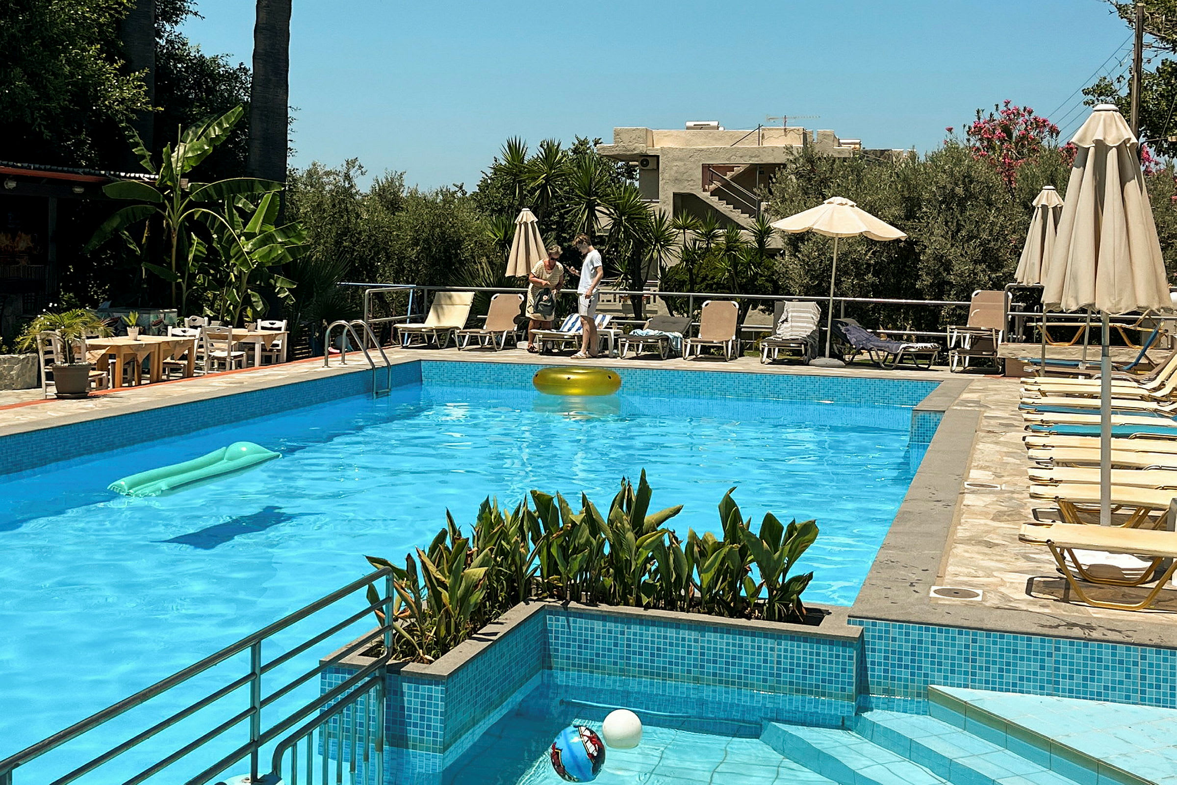 En swimmingpool med liggestole og parasoller omkring. I baggrunden ses beplantning og bygninger. (AI-genereret tekst)