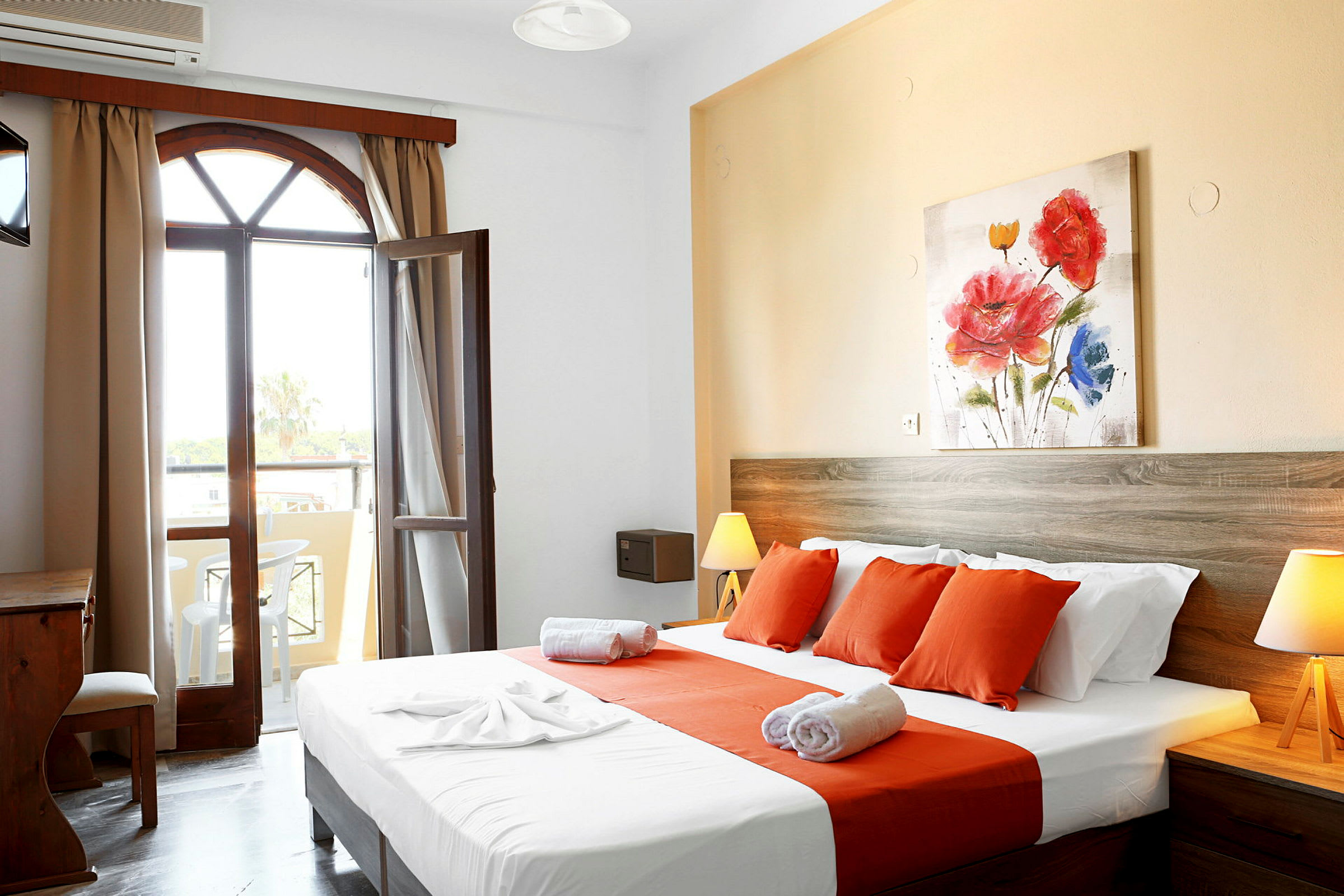 Et hotelværelse med en seng dækket med hvide og orange lagner, et træ-skrivebord og en åbning mod en balkon. (AI-genereret tekst)