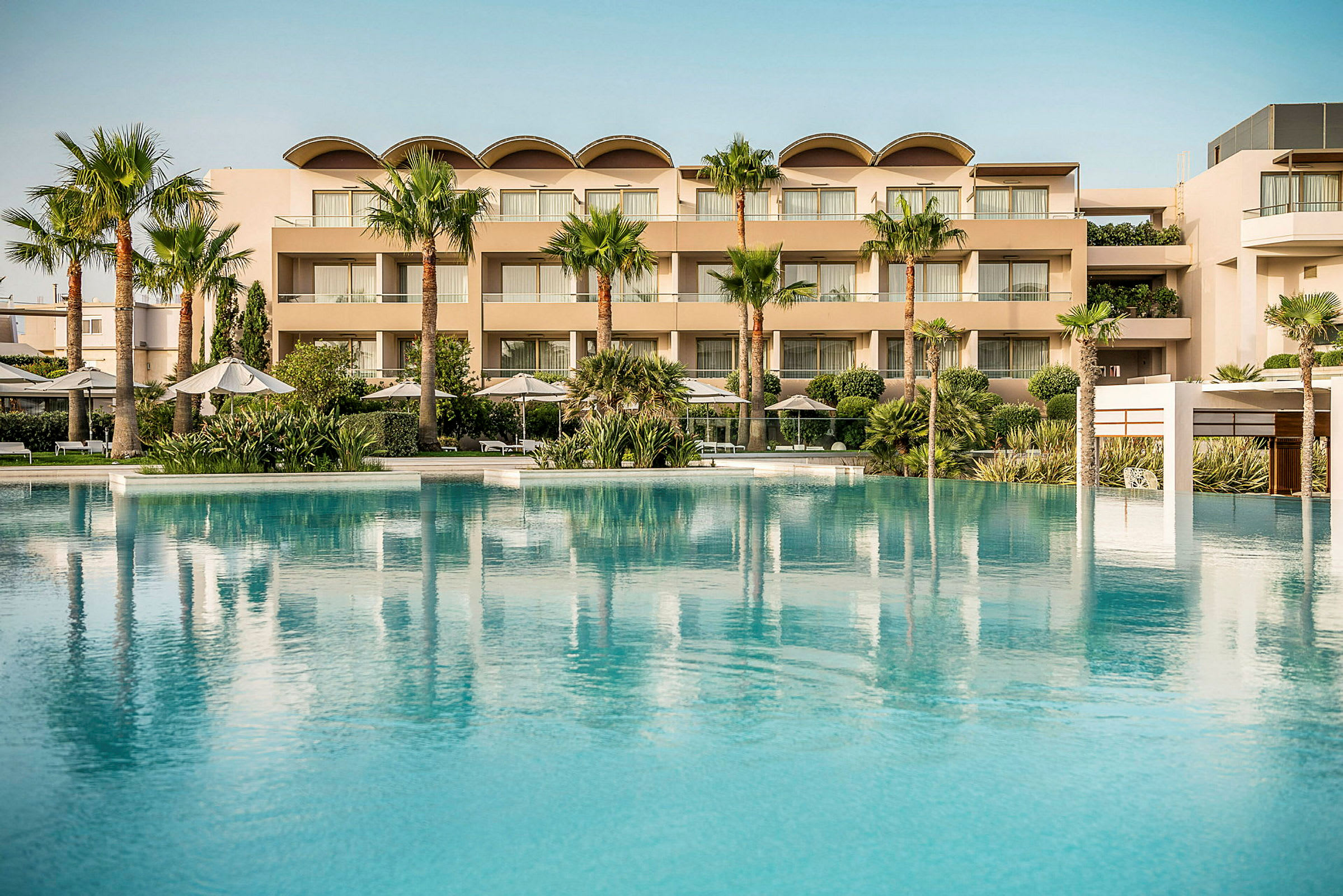 Et stort poolområde med klart blå vand foran et moderne hotel med flere etager og palmer samt beplantning. (AI-genereret tekst)