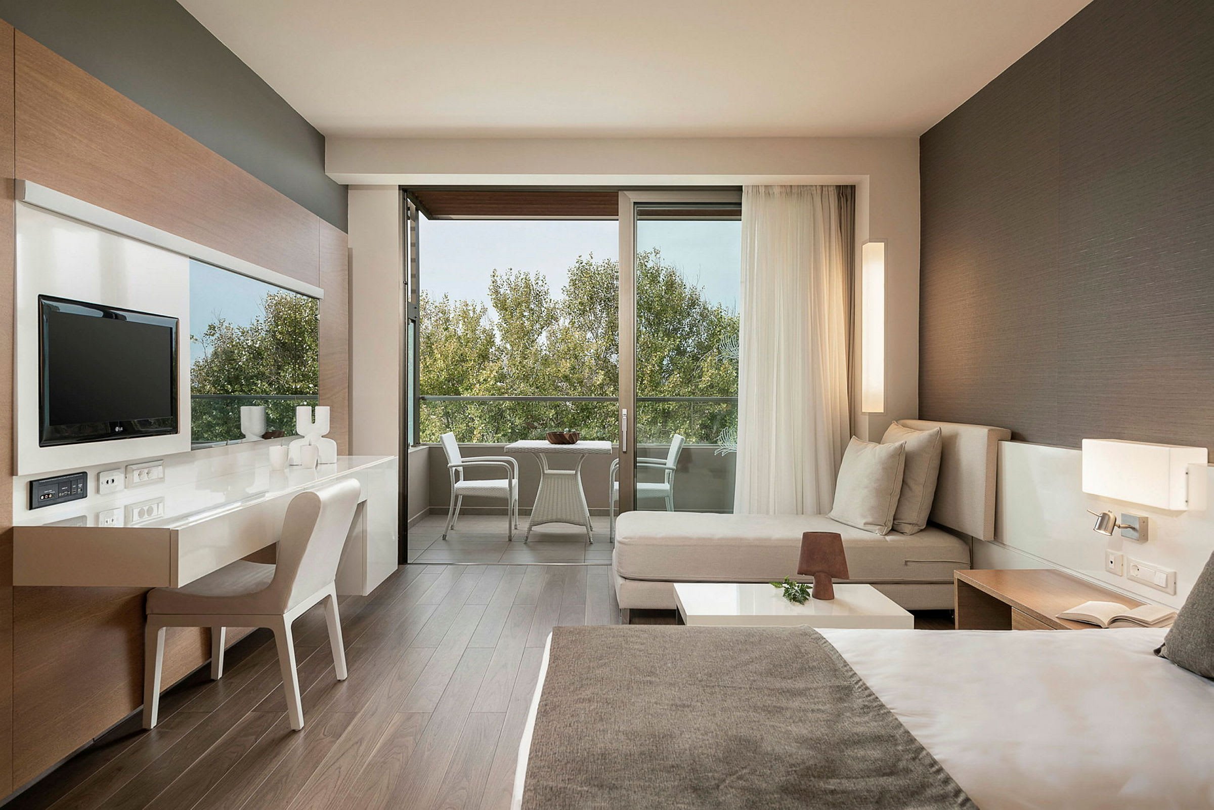 Et moderne hotelværelse med en dobbeltseng, siddeområde, skrivebord og et fjernsyn. Værelset har en stor balkon med havemøbler og udsigt til træer. (AI-genereret tekst)