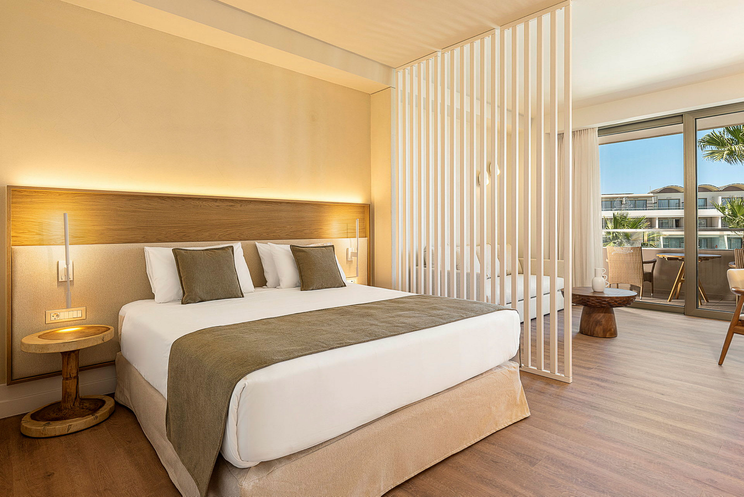 Et moderne hotelværelse med en dobbeltseng, dekorative puder og et lille natbord. Værelset har en skillevæg og et siddeområde med udsigt til en balkon. (AI-genereret tekst)