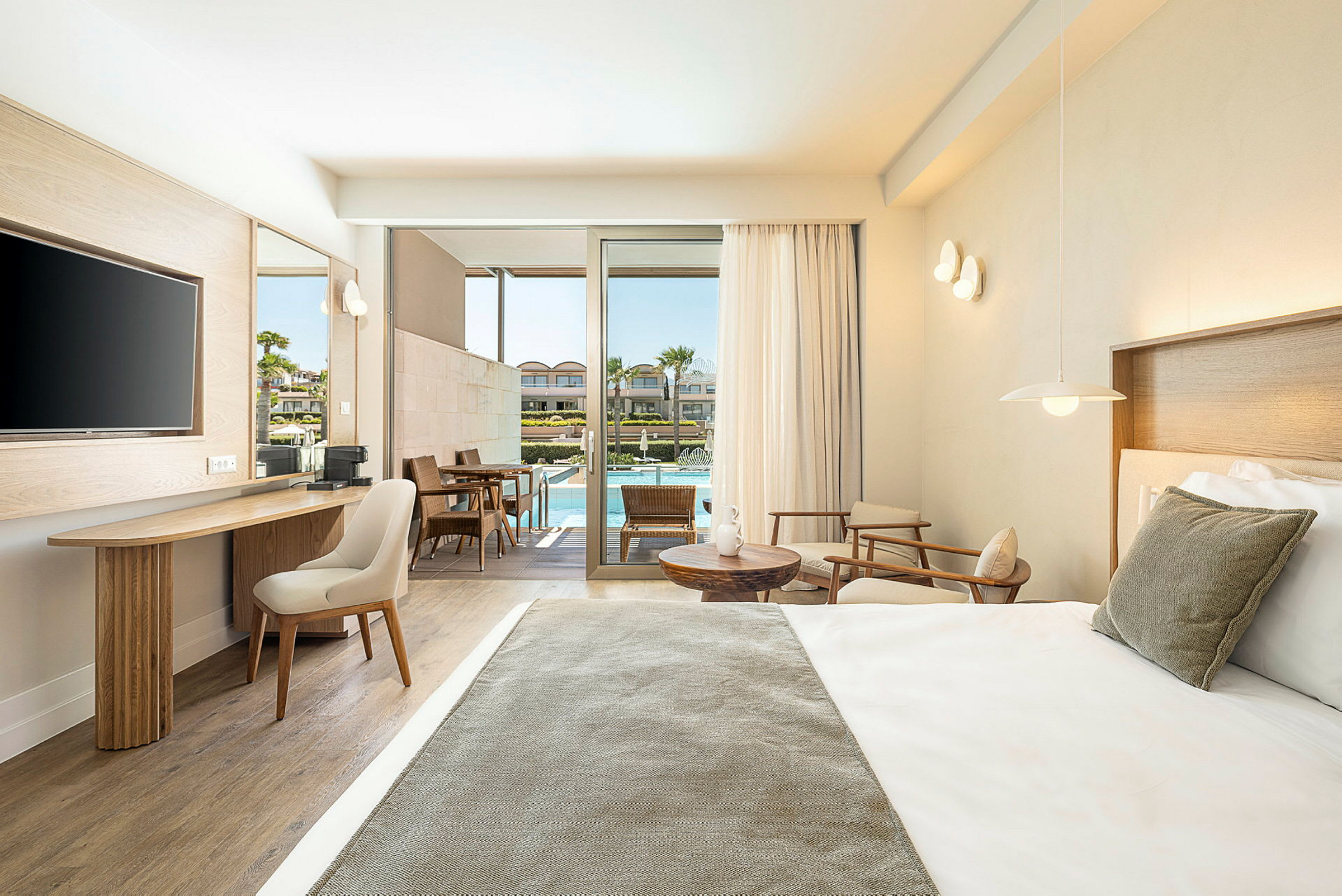 Et moderne hotelværelse med en redt seng, siddeområde og skrivebord. Værelset har store glasdøre ud til en terrasse med bord, stole og adgang til en pool. (AI-genereret tekst)