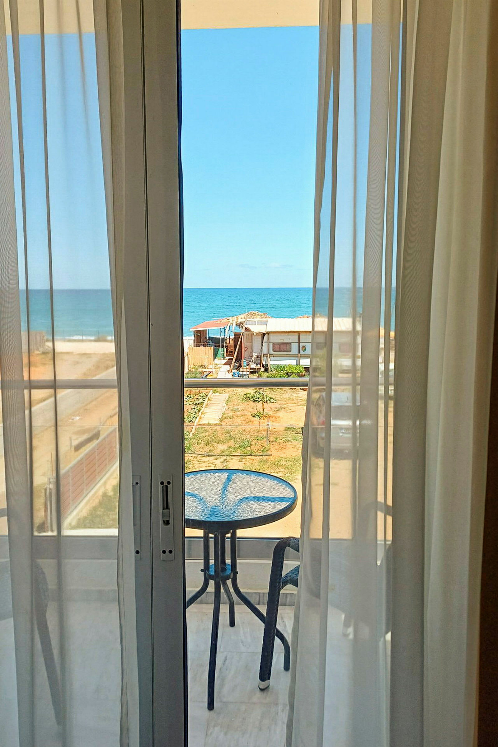 Udsigt gennem vindue med gardiner mod en balkon med et bord og stole samt havet i baggrunden. (AI-genereret tekst)