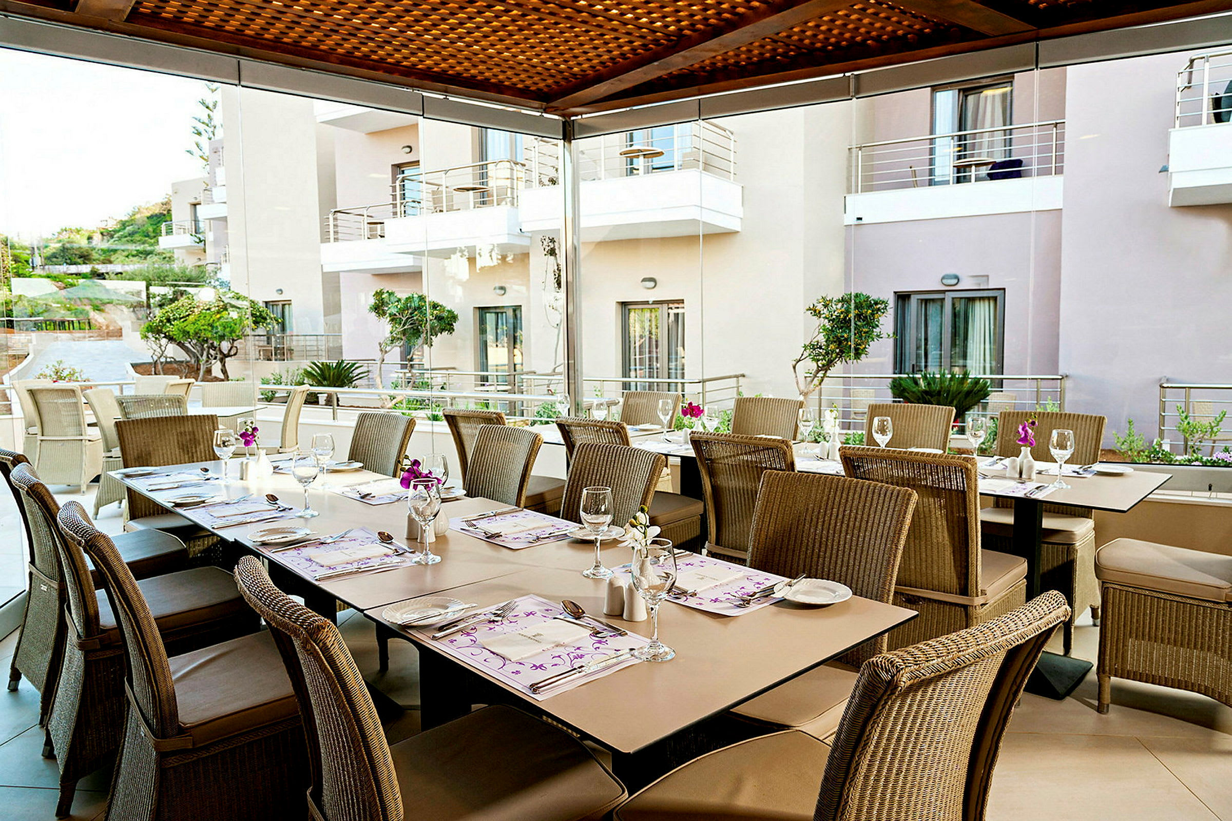 Borde og stole arrangeret på en terrasse med panoramavinduer, som giver udsigt til en bygning med altaner og grønne planter. (AI-genereret tekst)