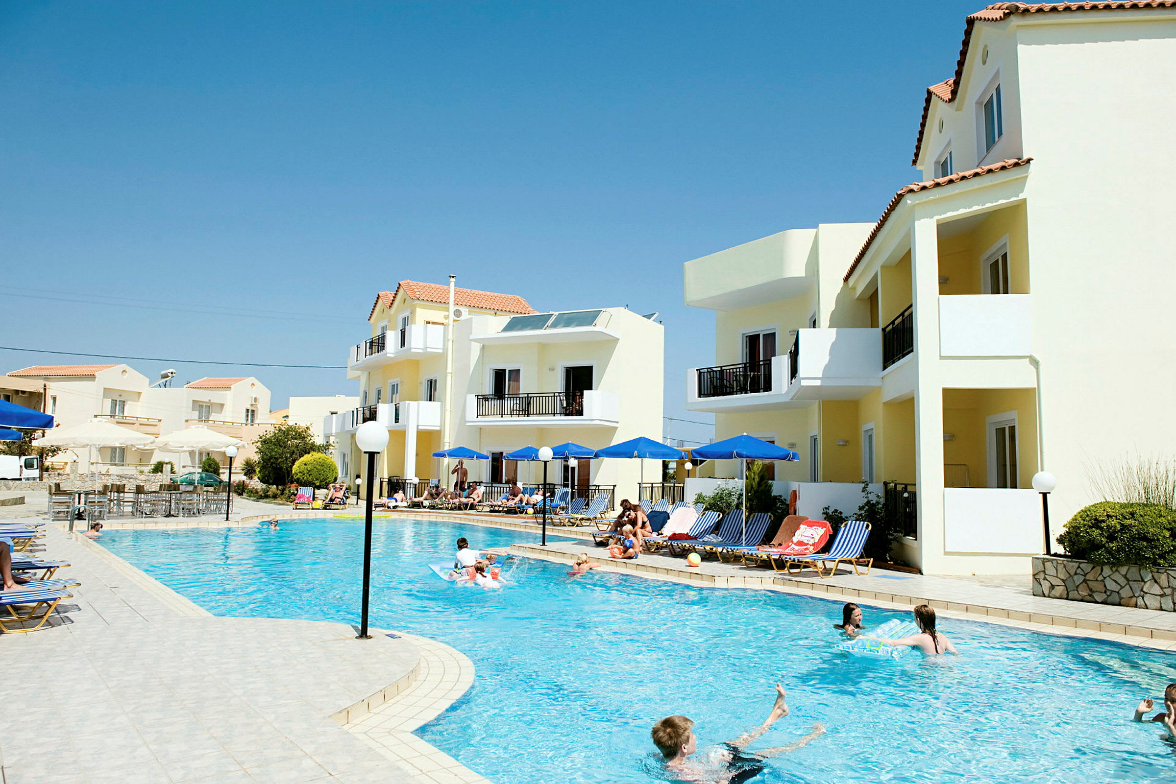 Udsigt over en pool med liggestole og parasoller ved siden, omgivet af lyse bygninger under en klar blå himmel. (AI-genereret tekst)