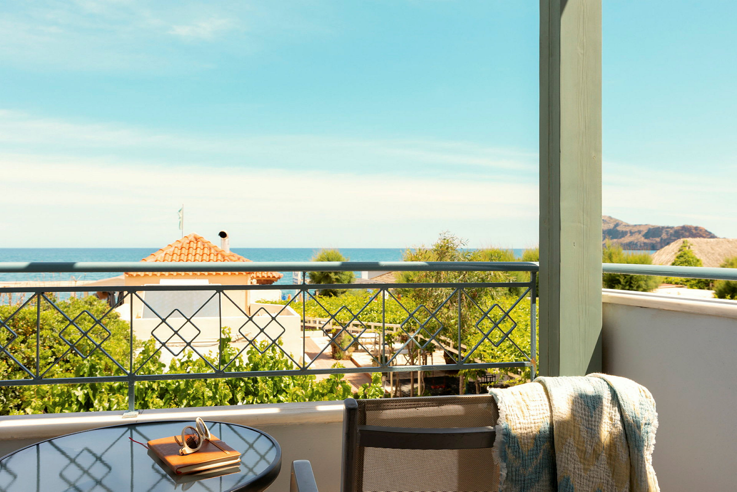 Udsigt fra en balkon med et glasbord og stol. Havet og huse ses i baggrunden. (AI-genereret tekst)