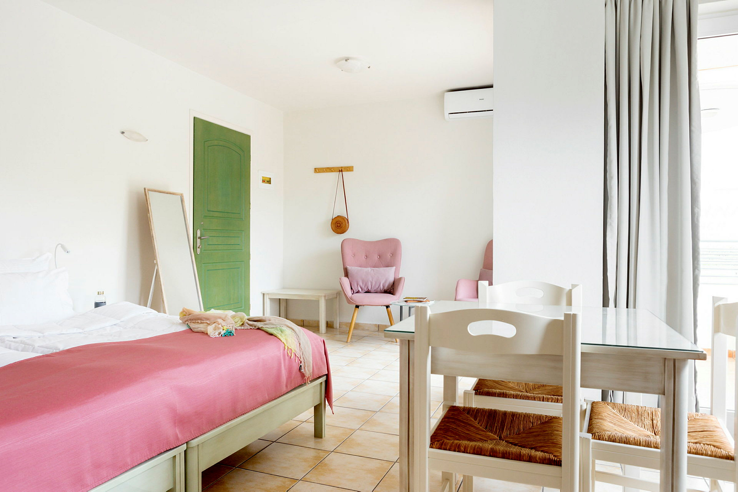 Et lyst og rummeligt hotelværelse med en grøn dør, en seng med rosa sengetæppe og et bord med stole. (AI-genereret tekst)