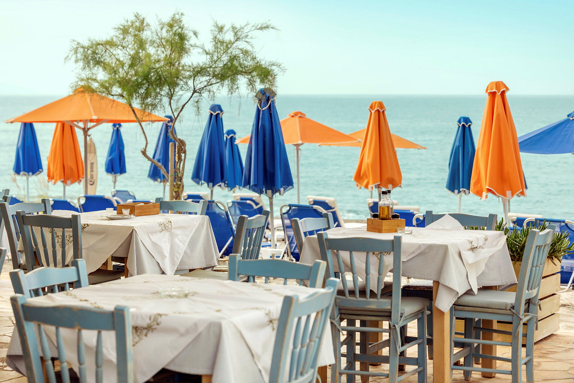 Et bord og stole står på en terrasse med udsigt over havet. I baggrunden ses parasoller i orange og blå på stranden. (AI-genereret tekst)