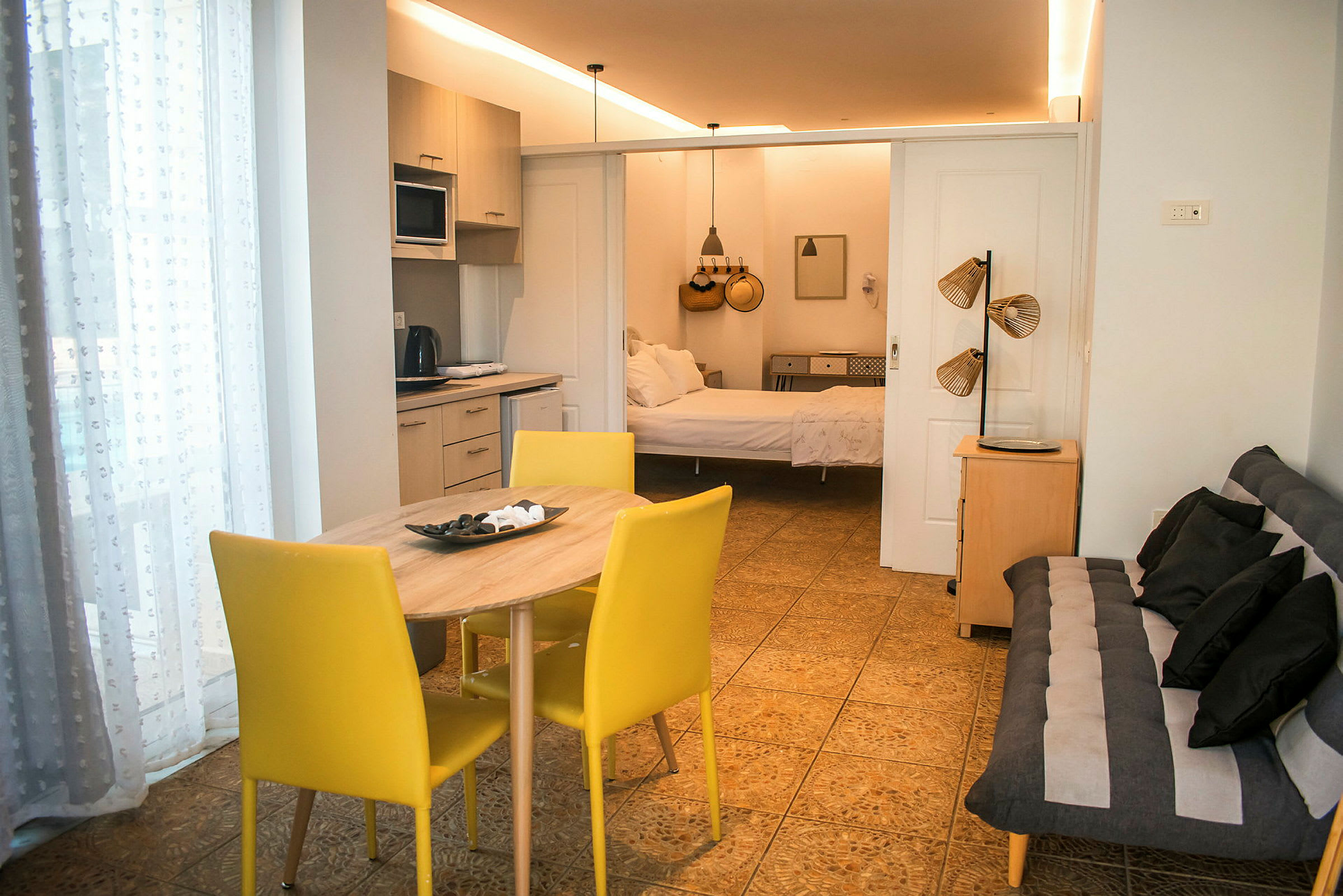 Interiør af en lejlighed med en kombineret stue og køkken samt en åben passage til soveværelset. Et spisebord med gule stole står i forgrunden. (AI-genereret tekst)