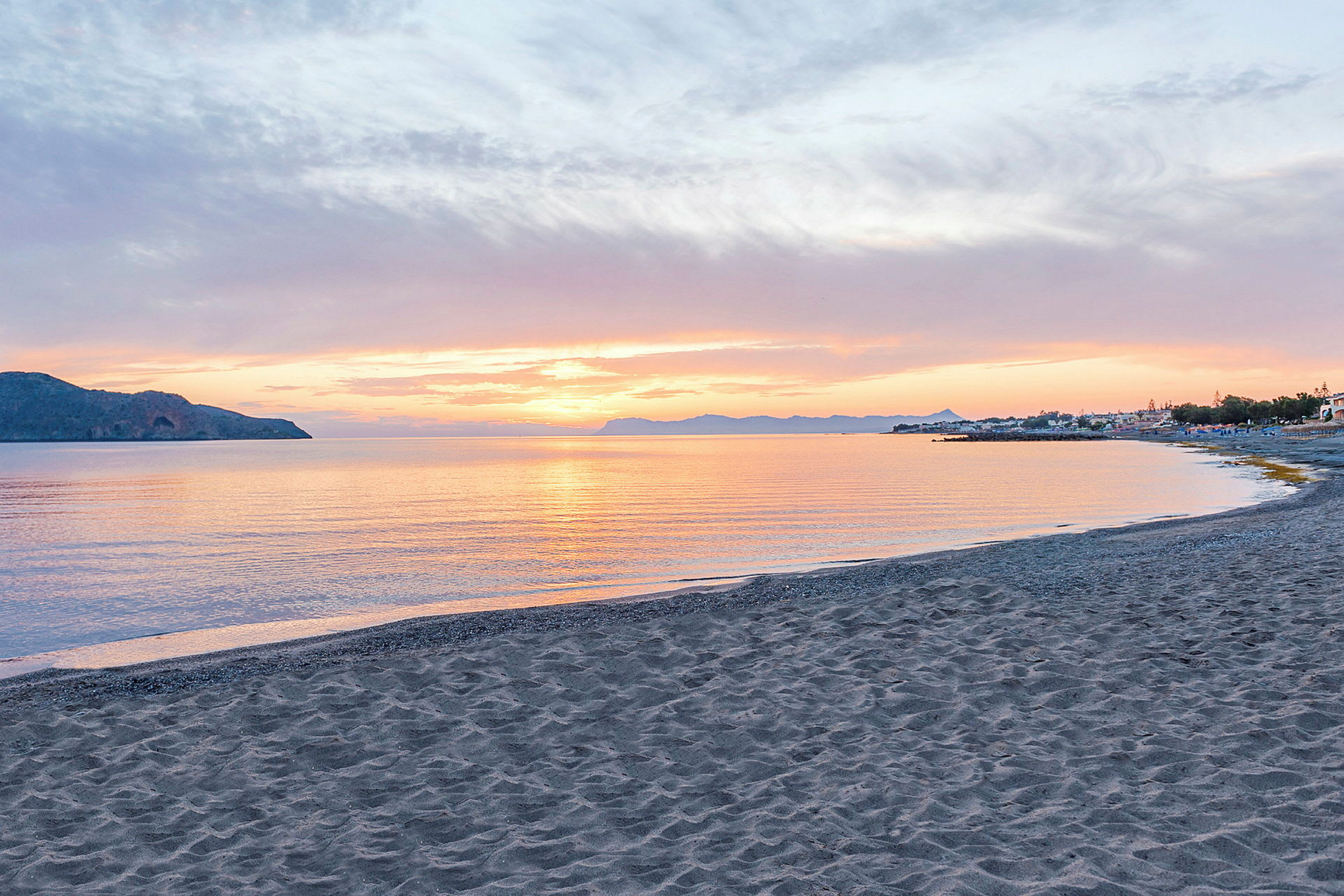 Solnedgang set fra en strand med havet i forgrunden og bjerge samt bygninger i baggrunden. (AI-genereret tekst)