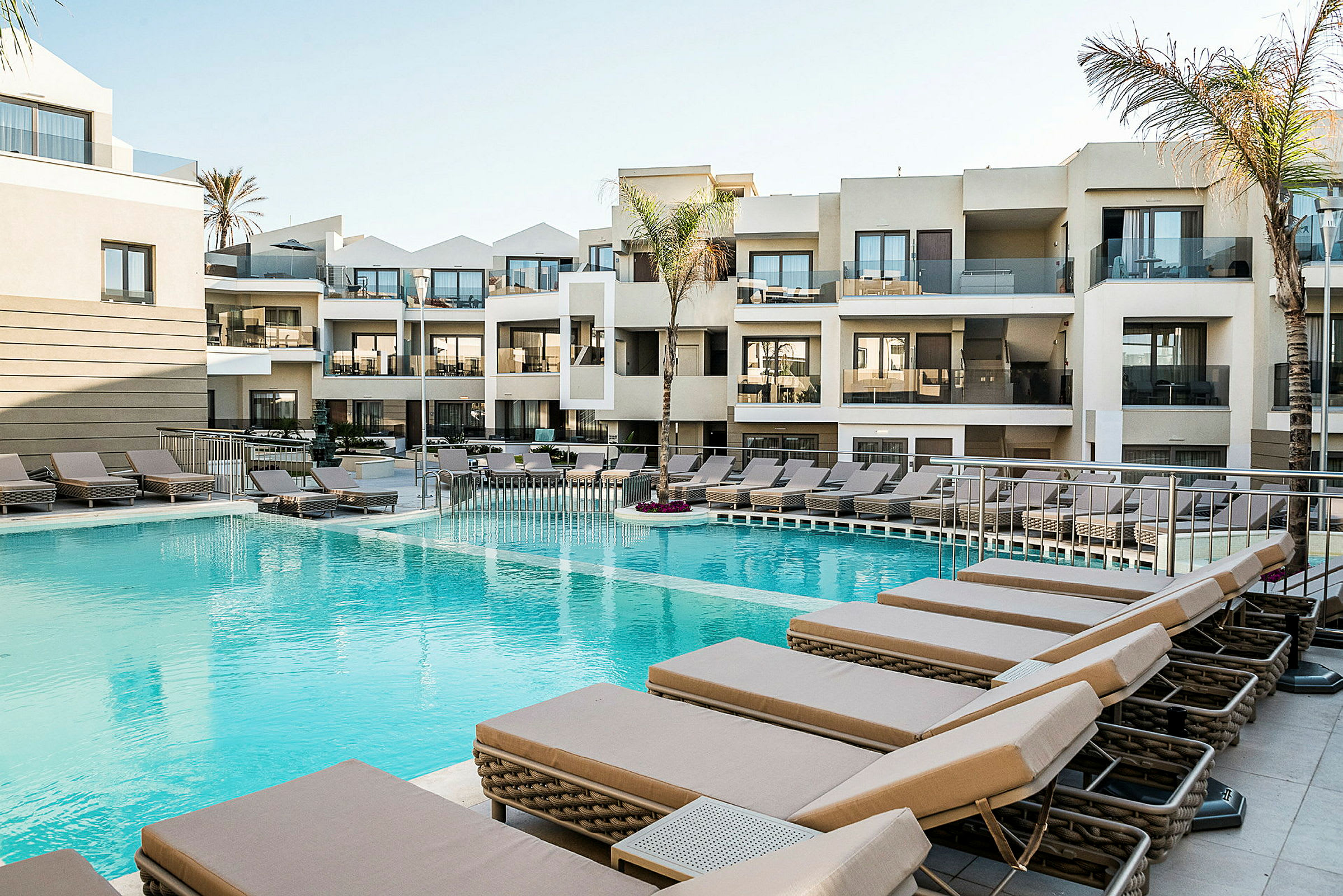 Hotelpool med liggestole og moderne bygninger i baggrunden. (AI-genereret tekst)