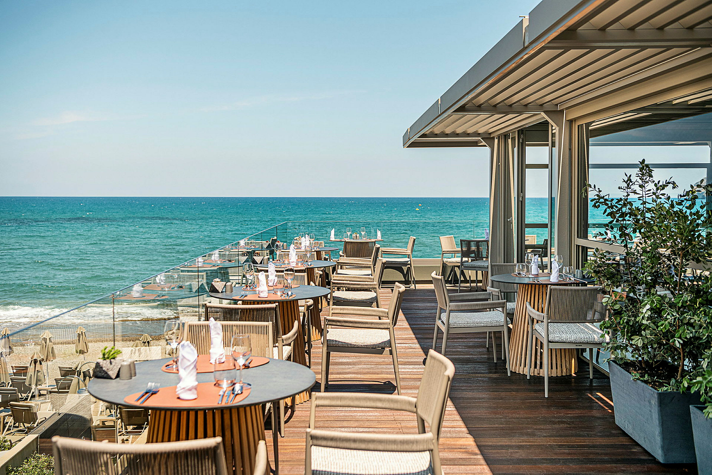 En terrasse med borde og stole ved havet. Blåt vand og klar himmel ses i baggrunden. (AI-genereret tekst)
