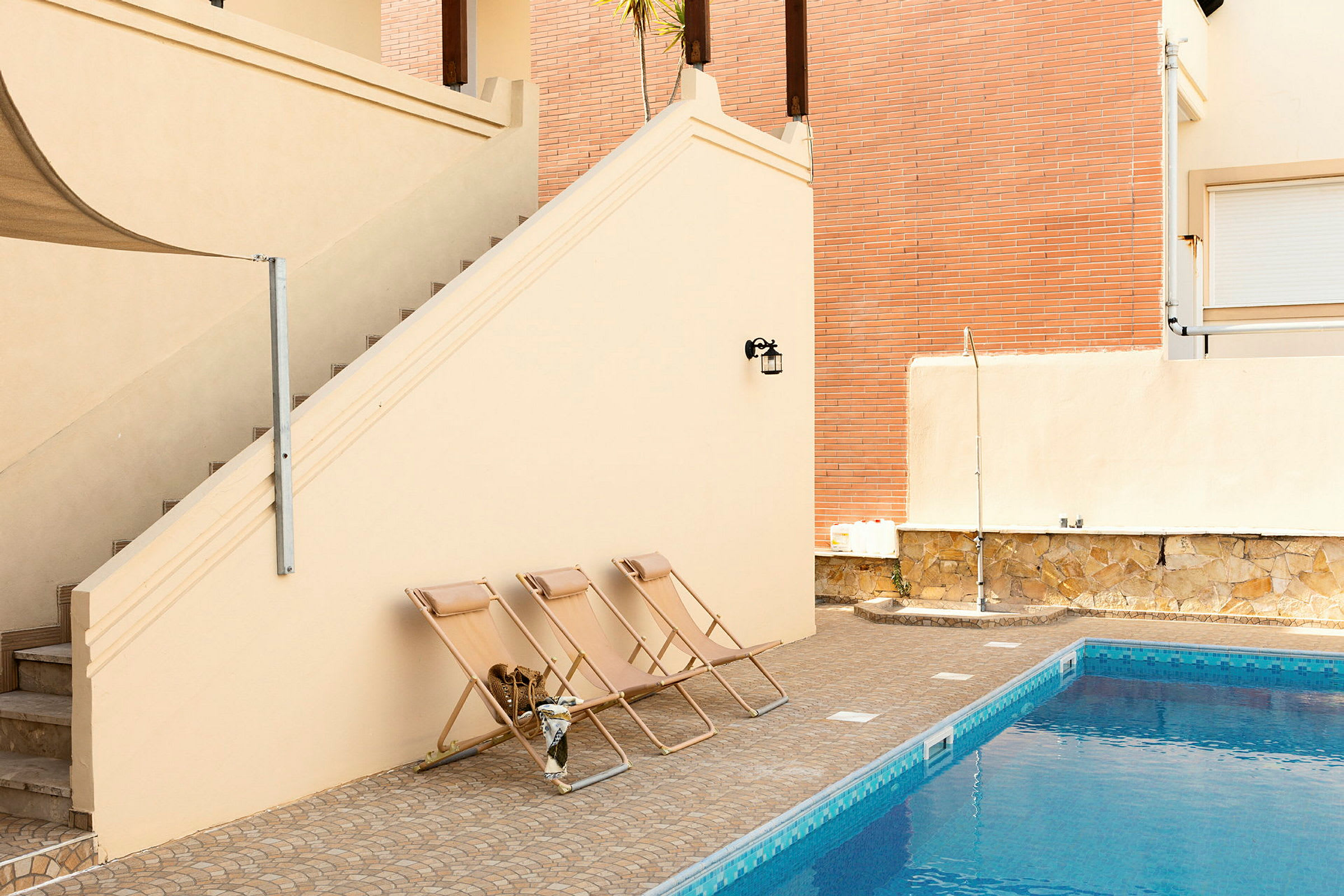Tre liggestole er placeret ved kanten af en swimmingpool. En trappe og facade udgør omgivelserne. (AI-genereret tekst)