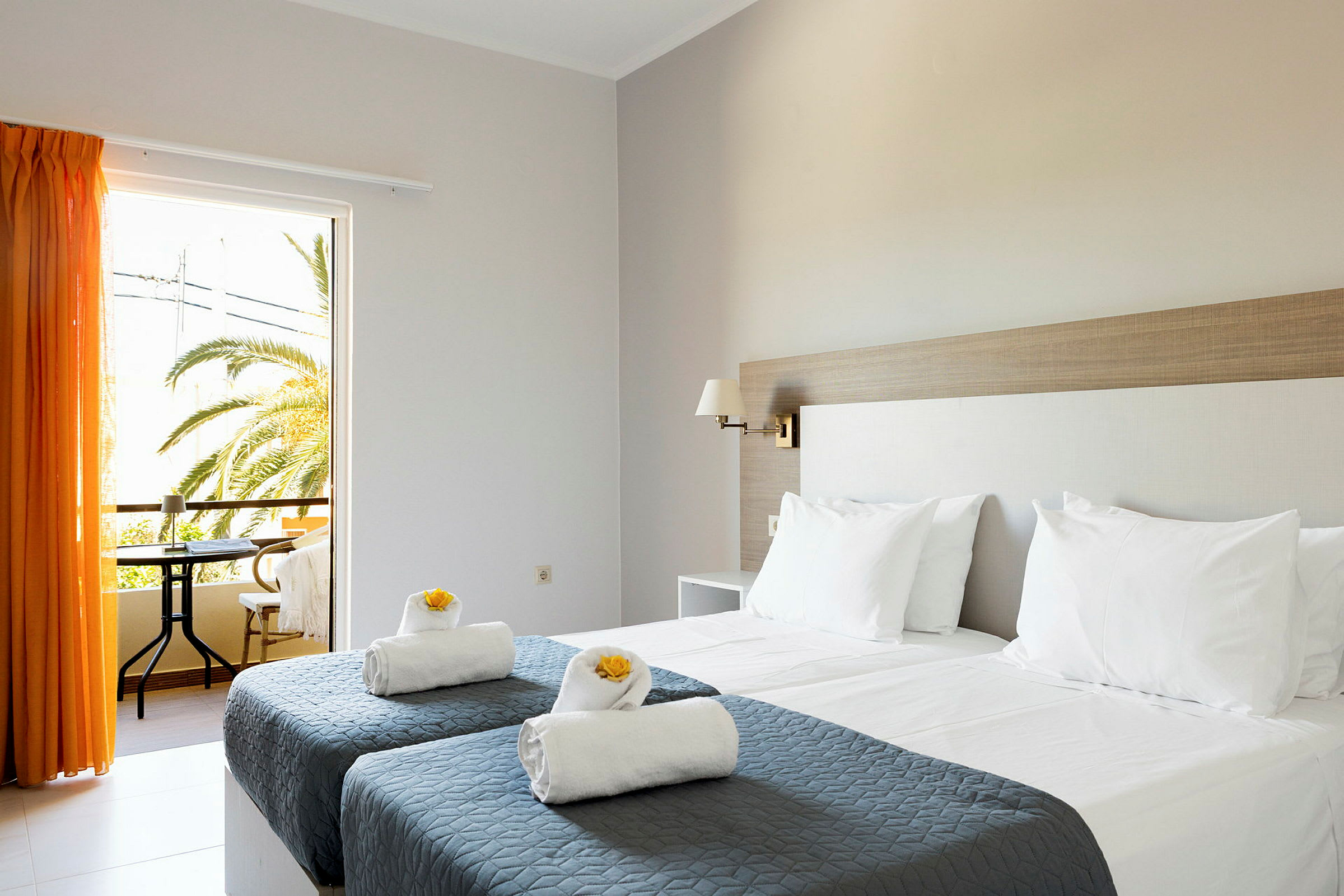 Et hotelværelse med to enkeltsenge med grå sengetæpper og hvide puder samt håndklæder dekoreret med gule blomster. Et vindue med orange gardiner åbner ud til en altan med udsigt over palmer. (AI-genereret tekst)