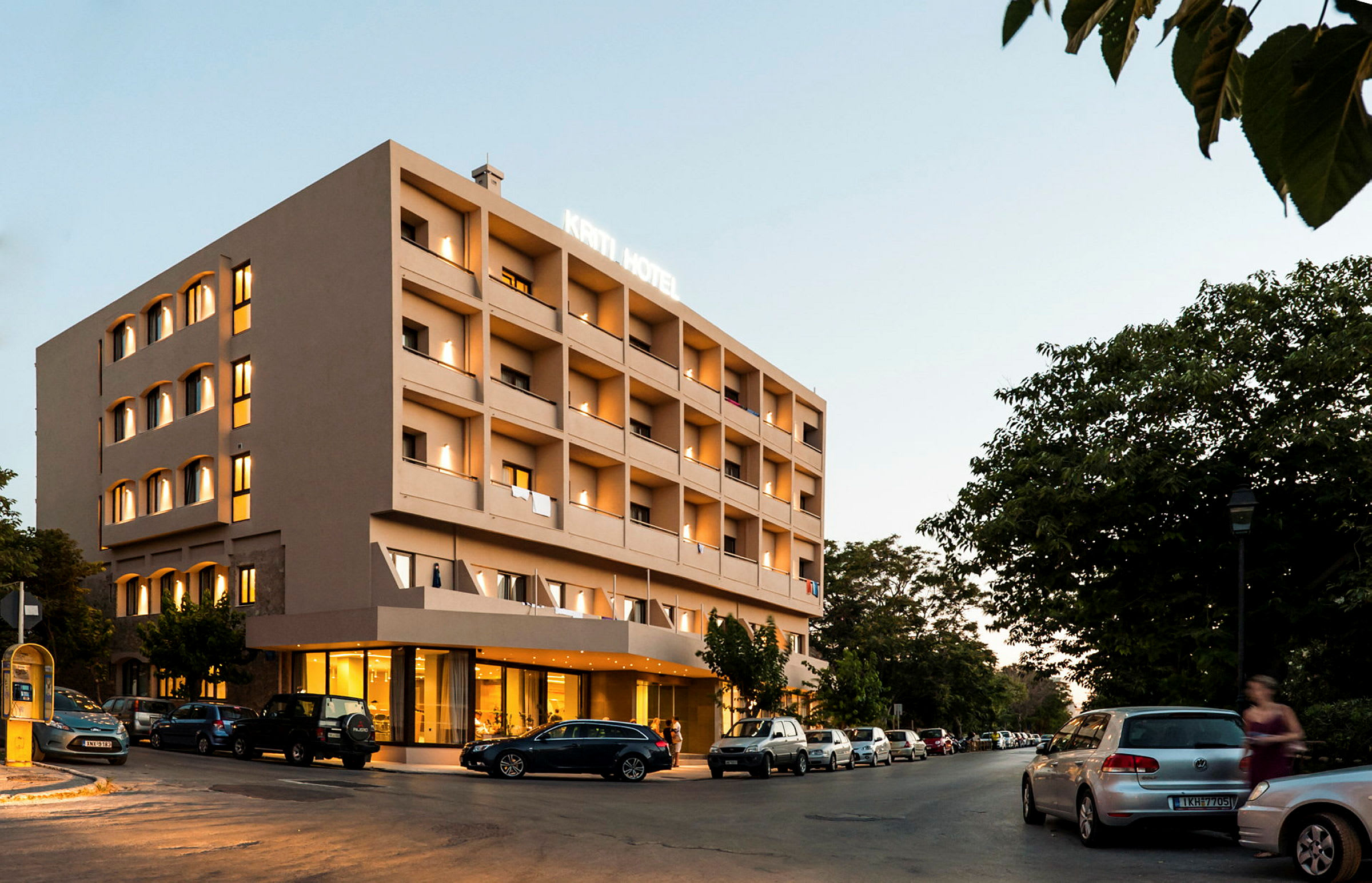 Et hotel med fire etager, oplyst facade, parkerede biler omkring og grønne træer i baggrunden. (AI-genereret tekst)